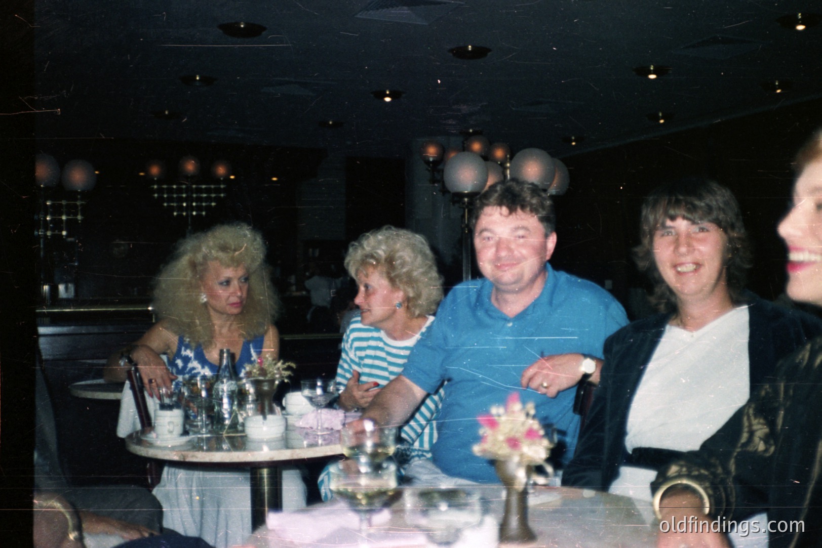 A social gathering at an upscale venue, likely a restaurant or private club. Four individuals are seated at a table featuring floral arrangements, glassware, and partial tableware. The style suggests the 1980s, indicated by hairstyles and clothing.