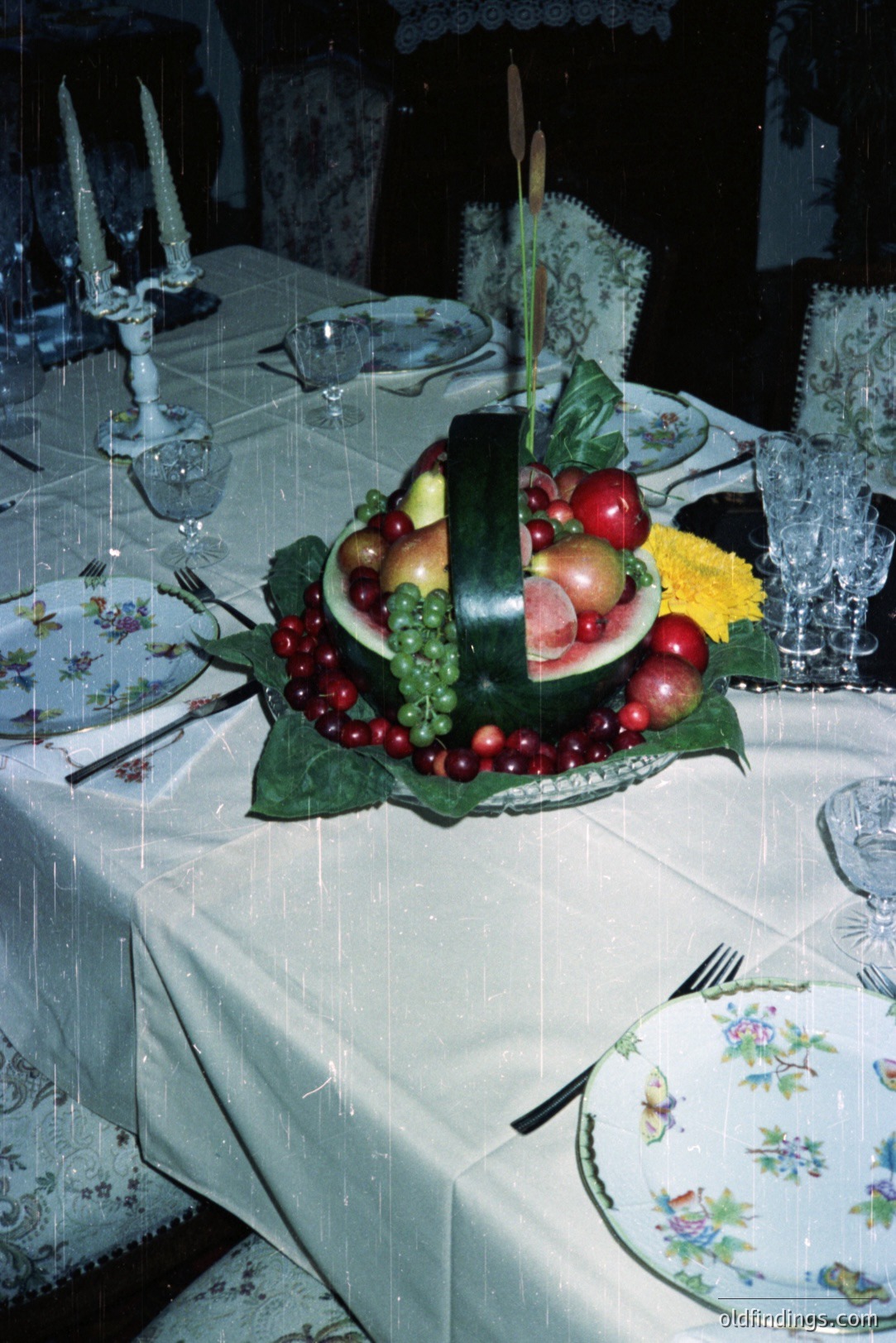 Elaborate centerpiece features a carved watermelon, displaying fruits like cherries, grapes, and apples on a bed of greenery. A floral patterned table cloth and vintage china set the scene, reminiscent of a formal dinner. Likely from the 1970s or 1980s.