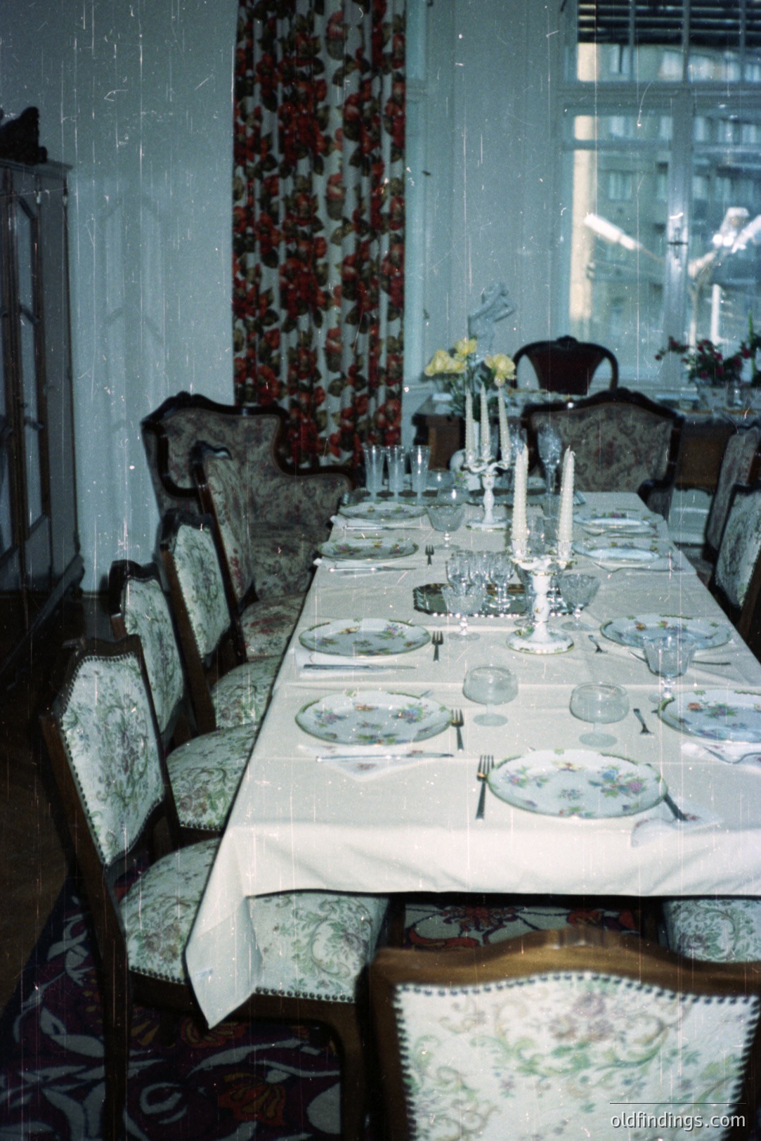 Elegant dining room scene: long table set for a formal gathering. Floral wallpaper, patterned chairs, and elaborate tableware suggest a 1970s aesthetic. Candle centerpiece and layered table linens indicate a special occasion. Appears to be European architecture.