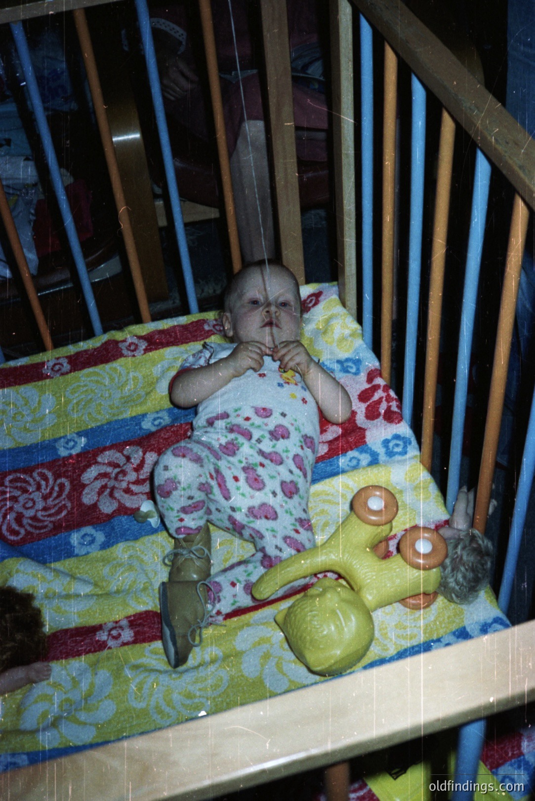 Infant in a crib, wearing patterned clothing, clutches a yellow plush toy. Crib bedding features a vibrant, geometric pattern. Photo’s faded colors and visible grain suggest a 1970s snapshot. Likely a personal or family archival image.