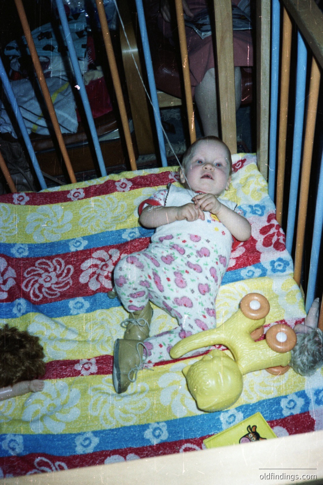 Infant seated in crib, wearing pink and white patterned clothing. Yellow rubber toy rests nearby. Appears to be a snapshot from the 1980s or 90s, typical of candid family photography. Crib design features blue and yellow detailing.