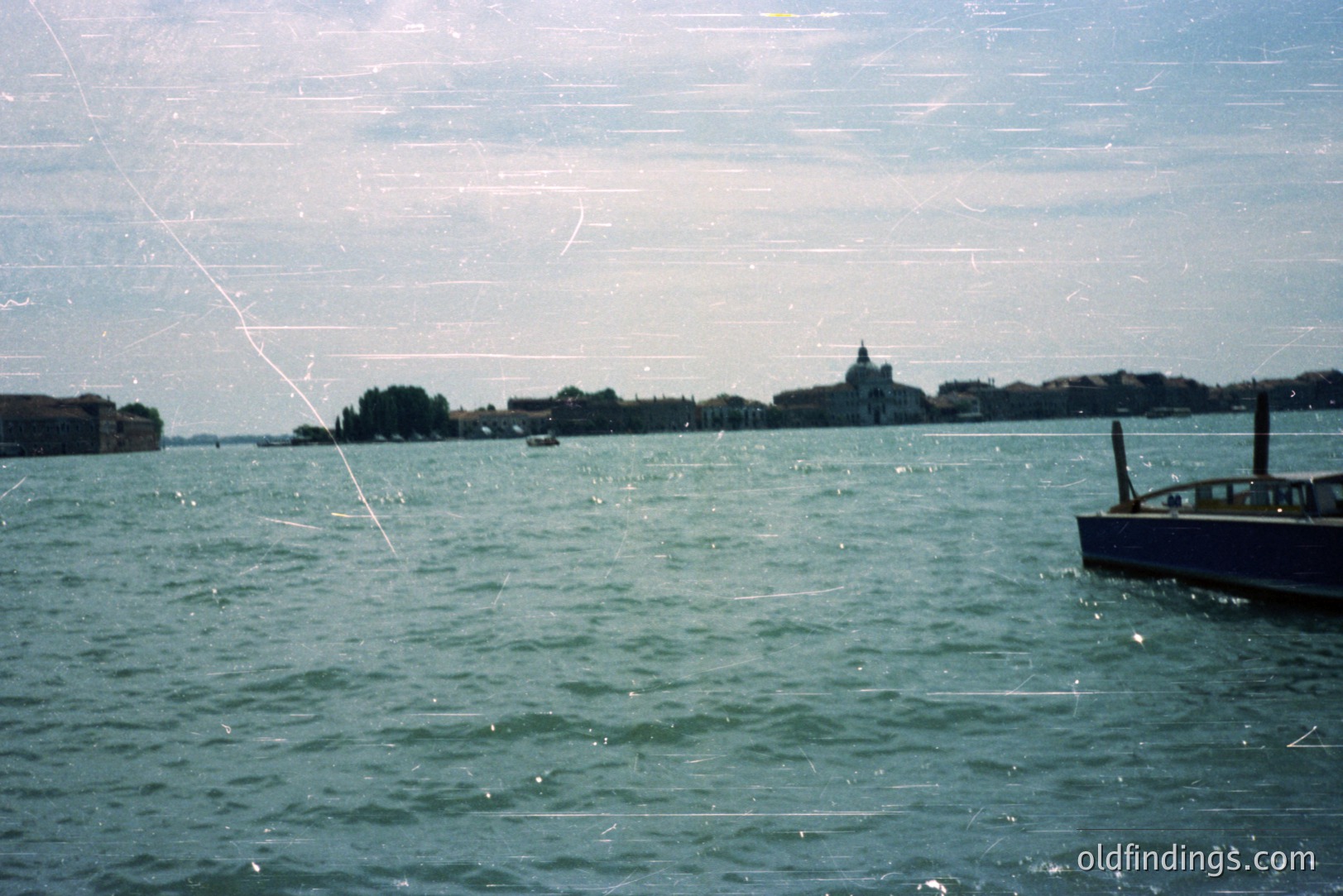 A view of the Venetian lagoon, featuring a classic wooden speedboat in the foreground and the island of San Giorgio Maggiore in the distance. The scene captures a hazy, atmospheric day. Likely taken in the 1970s or 80s, based on the film aesthetic and boat style. Potential stock imagery for travel or design.