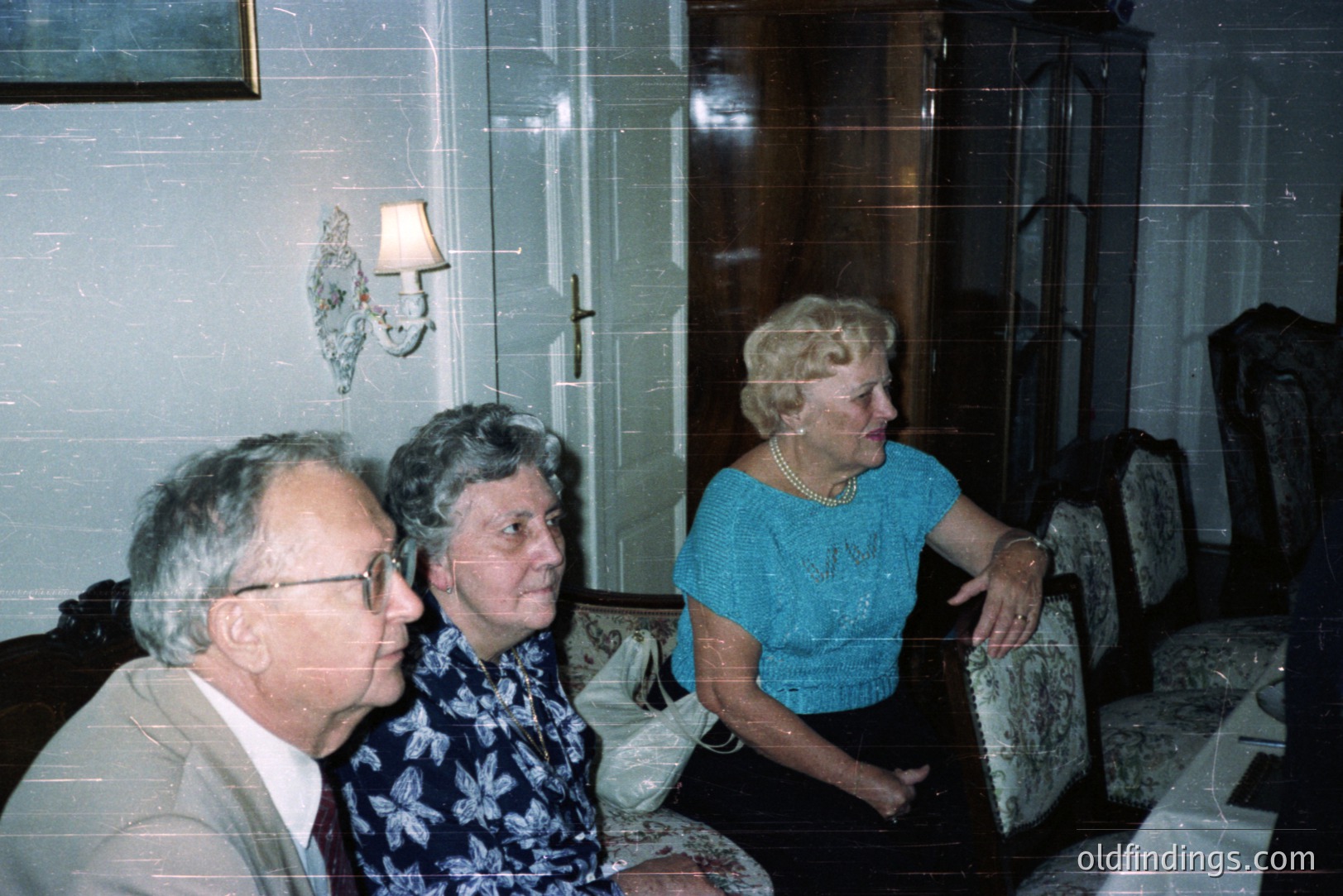 Three individuals seated in a formal living room, likely a family portrait. The man in a suit, two women in patterned blouses/tops. Antique furniture, wall sconce, and ornate wood detail define the decor. Likely taken in the 1970s or 80s. Possible design reference.