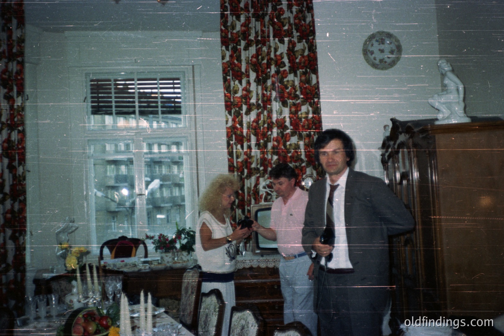Formal dining room interior with three figures. A woman adjusts a vase, a man in slacks stands, and another in a suit poses. Ornate floral wallpaper, tall windows, and a marble statue enhance the 1970s aesthetic. Likely residential, possibly a staged photograph.