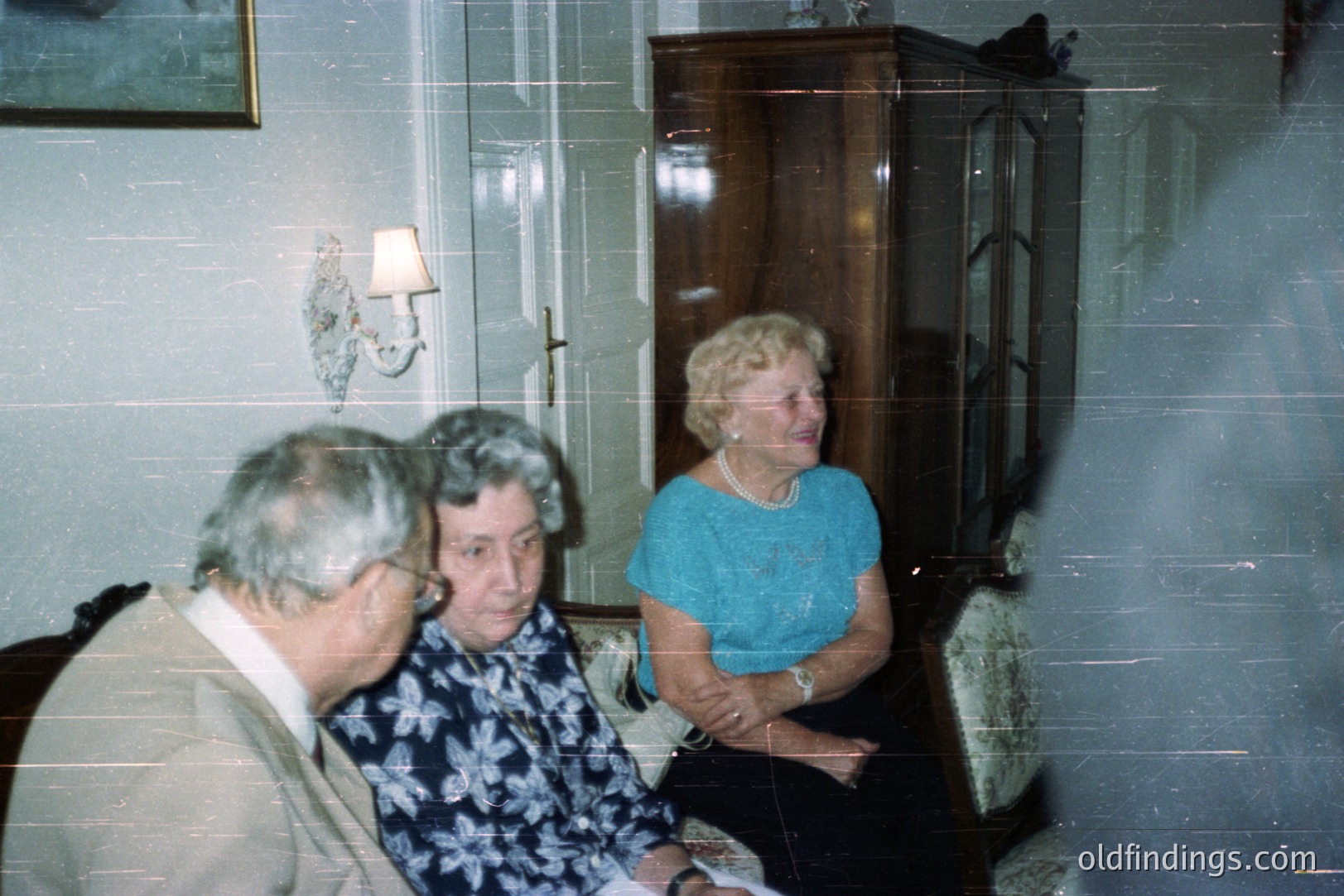 Three individuals sit on a patterned armchair in a well-lit room with a tall, dark wood cabinet and wall-mounted lamp. The man wears a suit jacket; women are in floral and teal tops. Appears to be a family portrait, likely 1970s or 80s. Warm color palette.