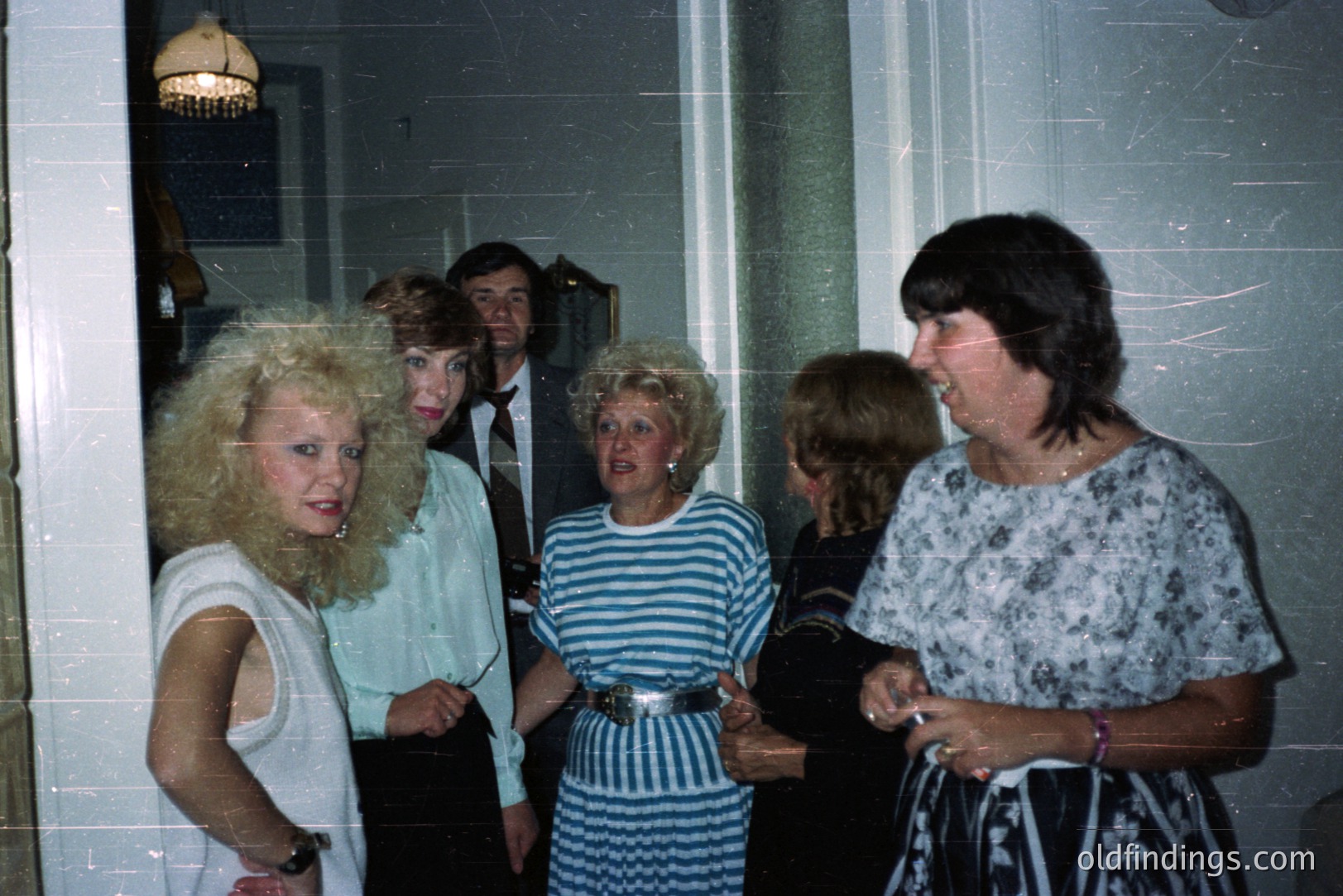 Candid moment: 5 people gather in a hallway, displaying 1970s fashion—large hair, wide collars, bold patterns. A man in a suit holds a camera. A floral dress and striped top are distinct. Likely a social gathering or event.