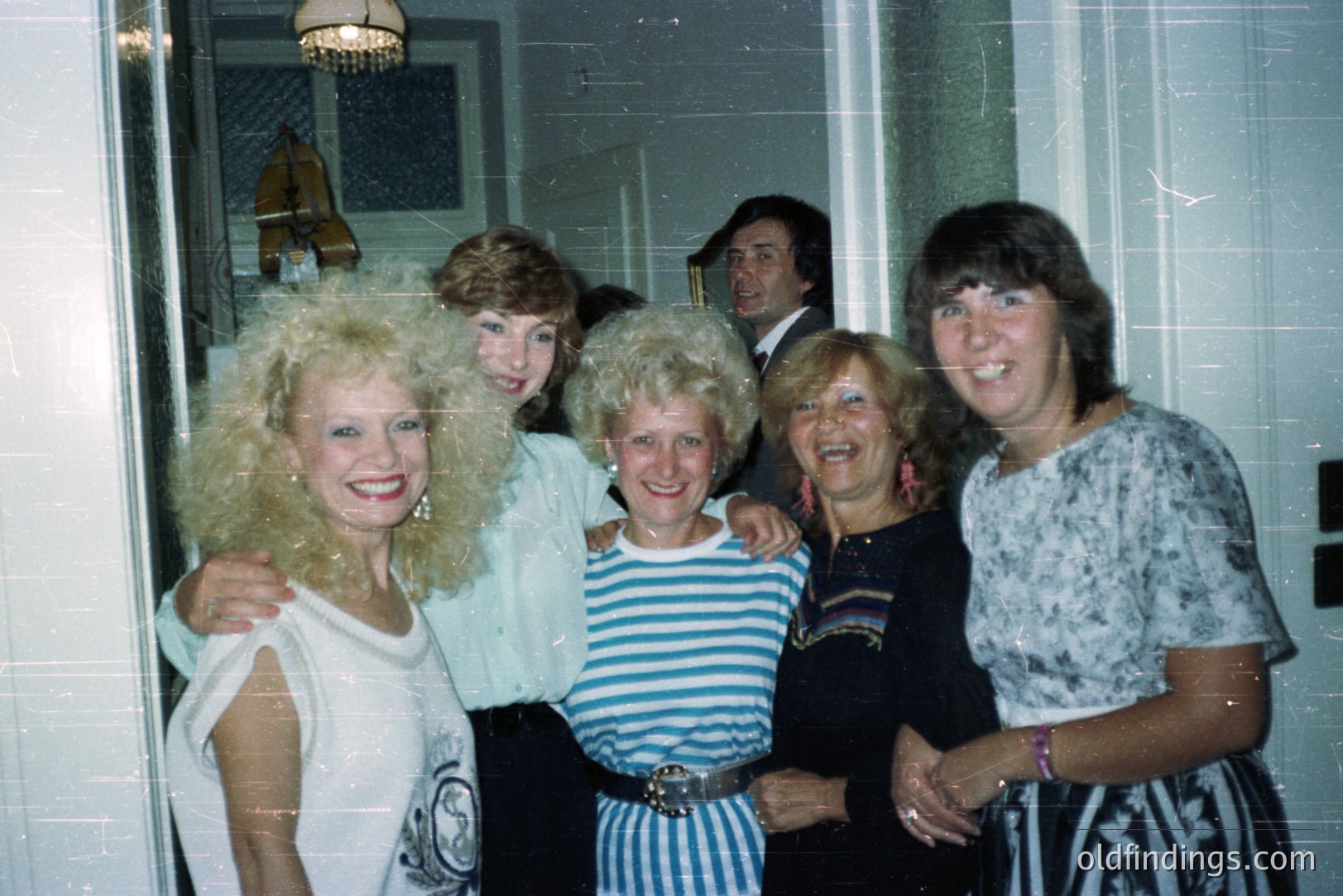 Group portrait of five people with voluminous hairstyles, characteristic of the late 1970s or early 1980s. A man stands partially visible behind the women. Interior setting with a patterned carpet & decorative lamps. Potential for stock photography referencing retro fashion.