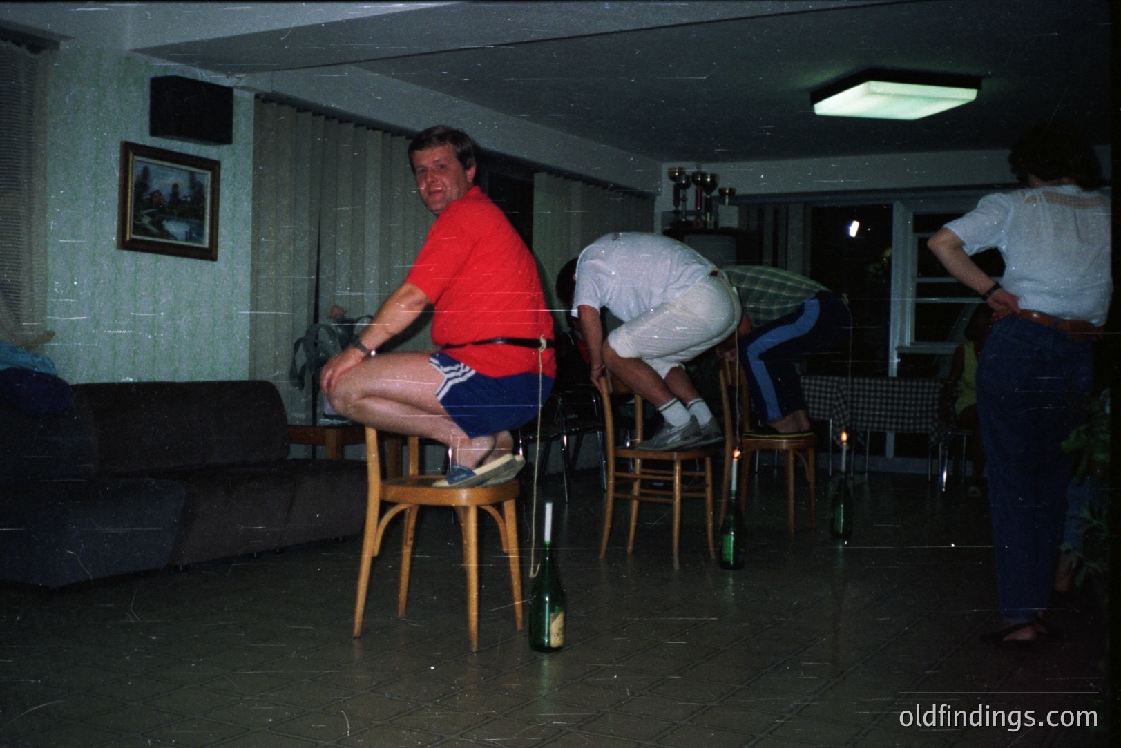 A staged scene featuring three men. One, wearing a red shirt and blue shorts, balances on a chair. Two other men are posed behind, attempting to dislodge the chair. Mid-century modern interior with patterned wallpaper & tile flooring. Likely 1970s, candid snapshot aesthetic.
