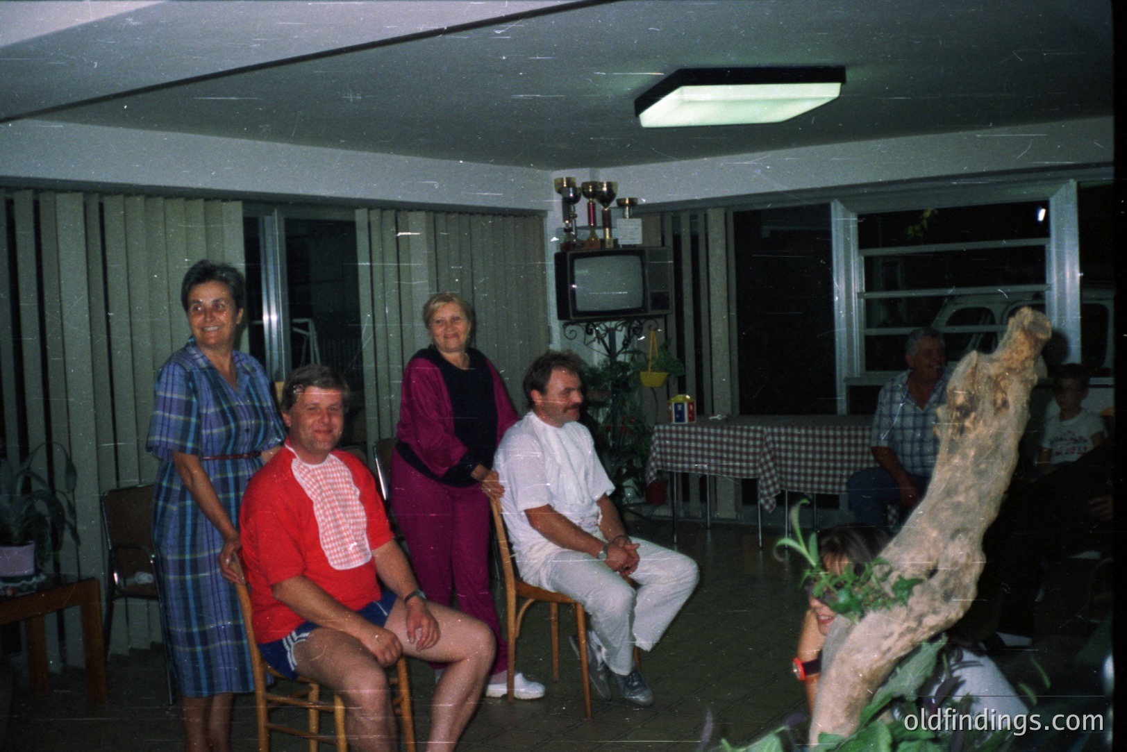 Group portrait inside a mid-century modern home, possibly a vacation rental. Five individuals pose; floral wallpaper and large windows are visible. Interior decor suggests the 1970s, likely central Europe. Trophy and a television set are displayed. A fish tank with a prominent branch dominates one corner.