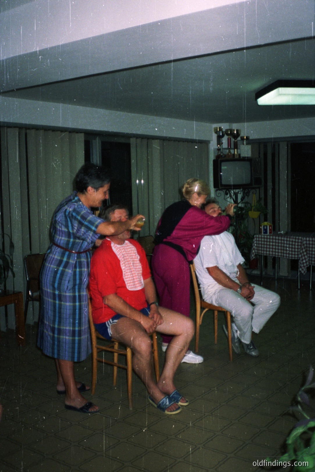Three figures inside an apartment appear to be experiencing a water leak, possibly a sprinkler malfunction. Two women in patterned robes are seemingly attempting to mitigate the spray. The men are seated; one in a red and white patterned shirt and swim trunks, the other in a white suit. Mid-century modern furnishings are visible.