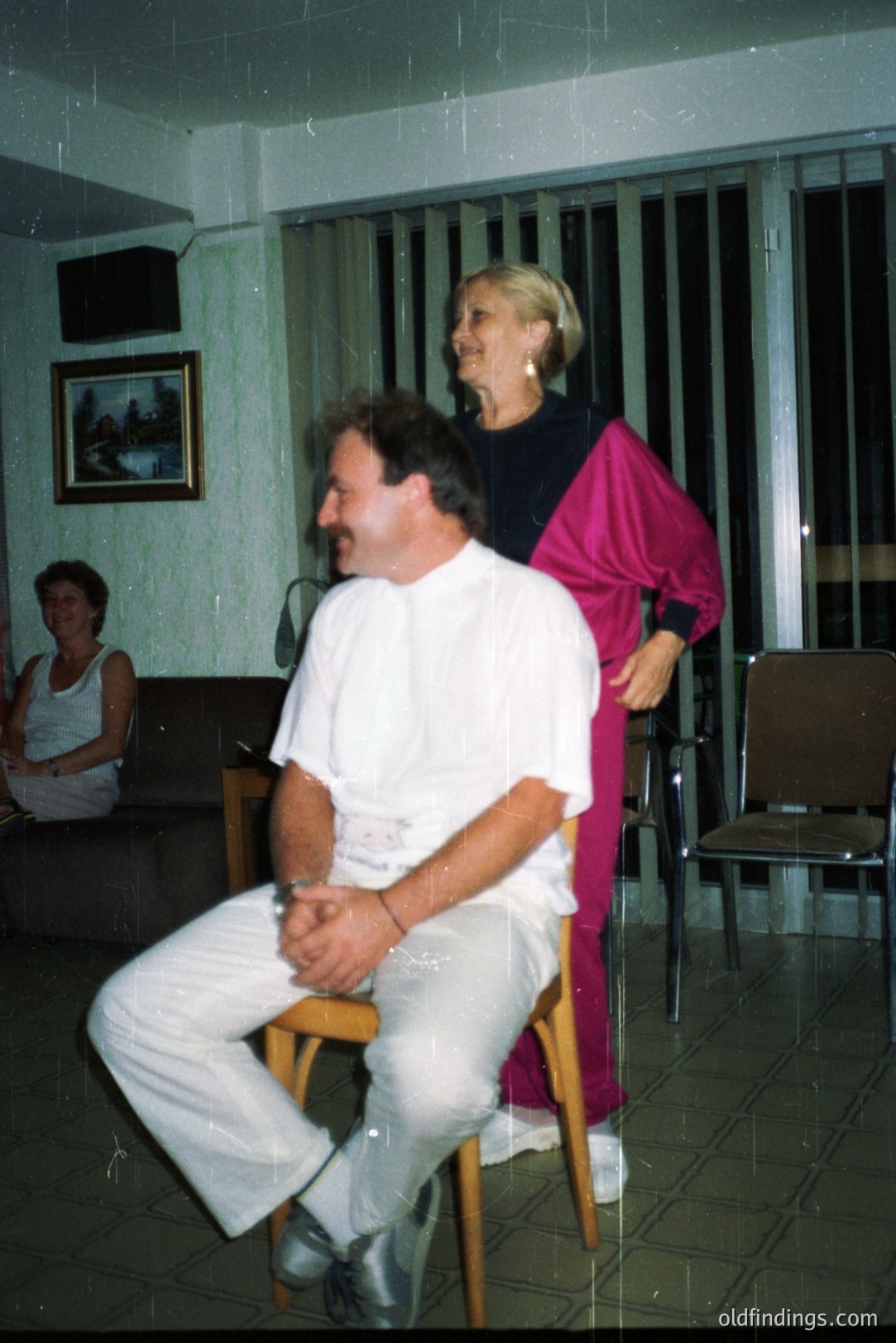 Man sits on a chair, smiling, with a woman in a pink tracksuit adjusting his collar. Interior scene, likely a recreation or social club, with tiled floor & blinds. Background shows a seated person and a framed print. Appears to be 1980s casual style.