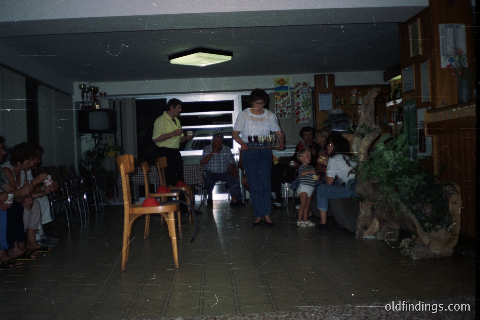 Interior scene depicts a 1970s-era hotel lobby or function room. A server carries drinks past seated guests and a TV console. Note distinctive wood-paneled walls and patterned tile flooring. Commercial potential for design reference/nostalgia stock.