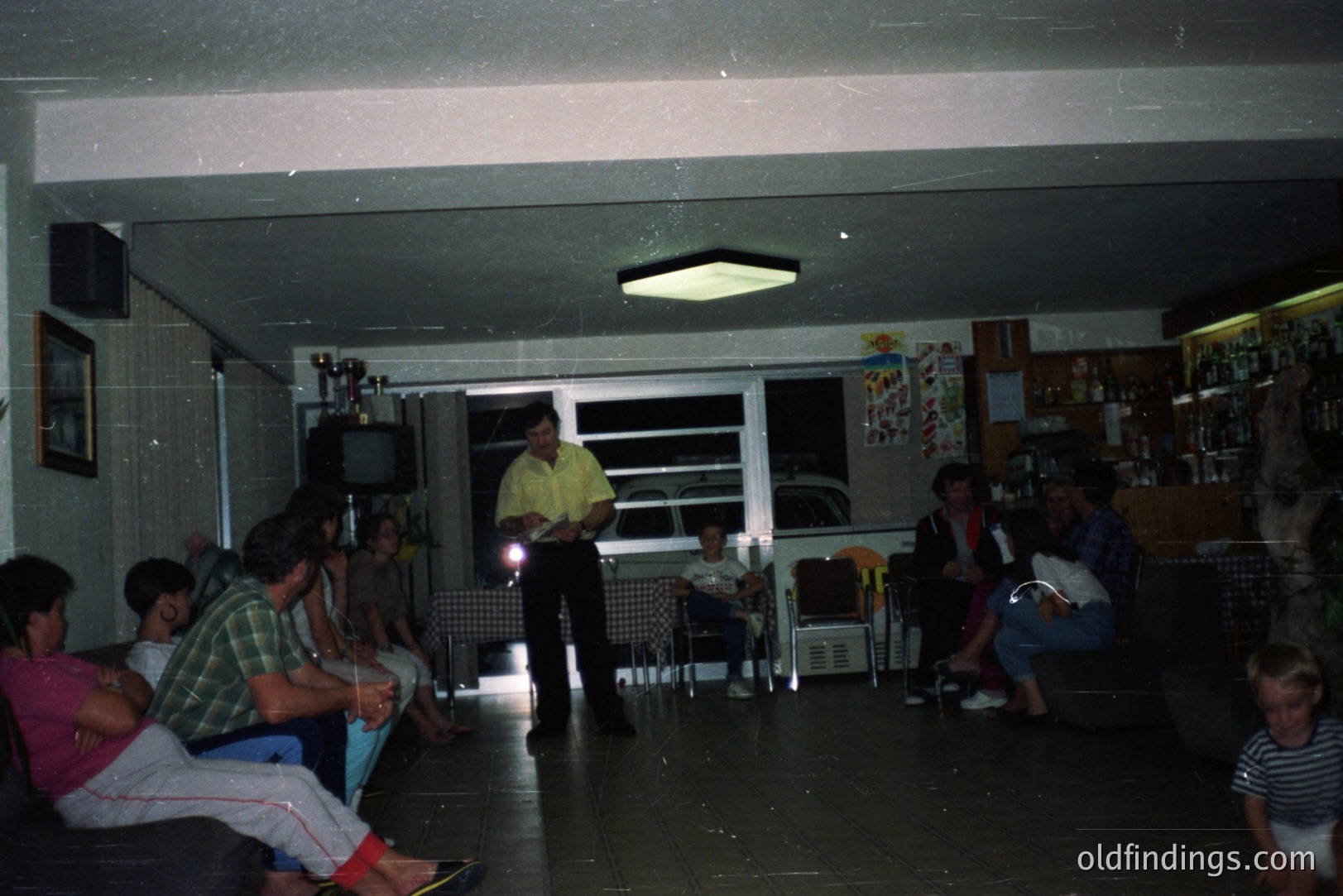A group gathers in a sparsely furnished room, likely a recreational space, featuring a television, trophy display, and a wet bar stocked with bottles. A man in a yellow shirt addresses the audience. The decor evokes the 1970s/80s, possibly a vacation home or community center.