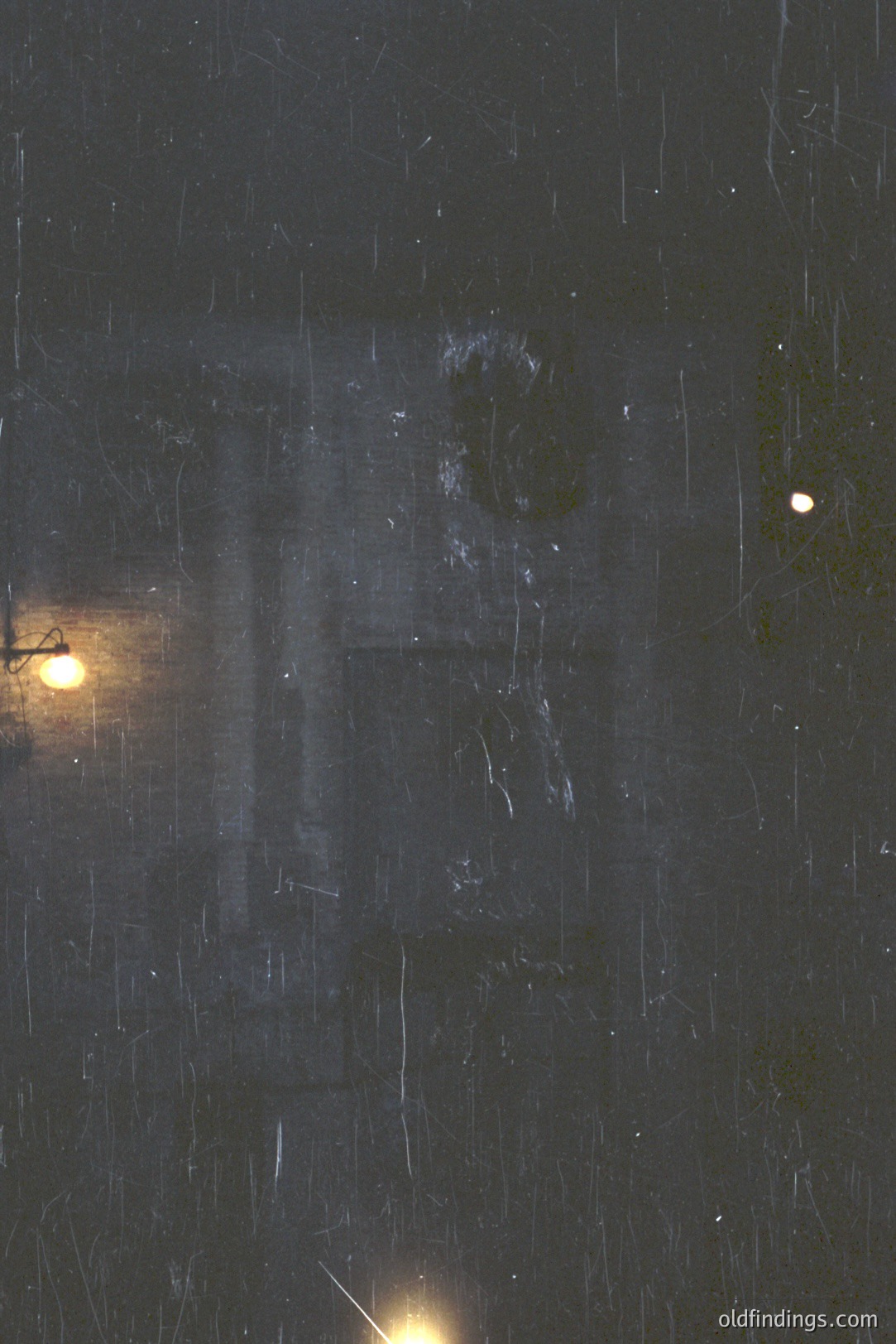 A blurred, rain-streaked exterior shot. A dimly lit doorway with a visible figure inside is partially obscured by water droplets. A vintage-style lamp glows nearby; a single light is visible above. Likely captured in a humid climate, possibly urban setting. Could be stock photo for atmospheric mood.