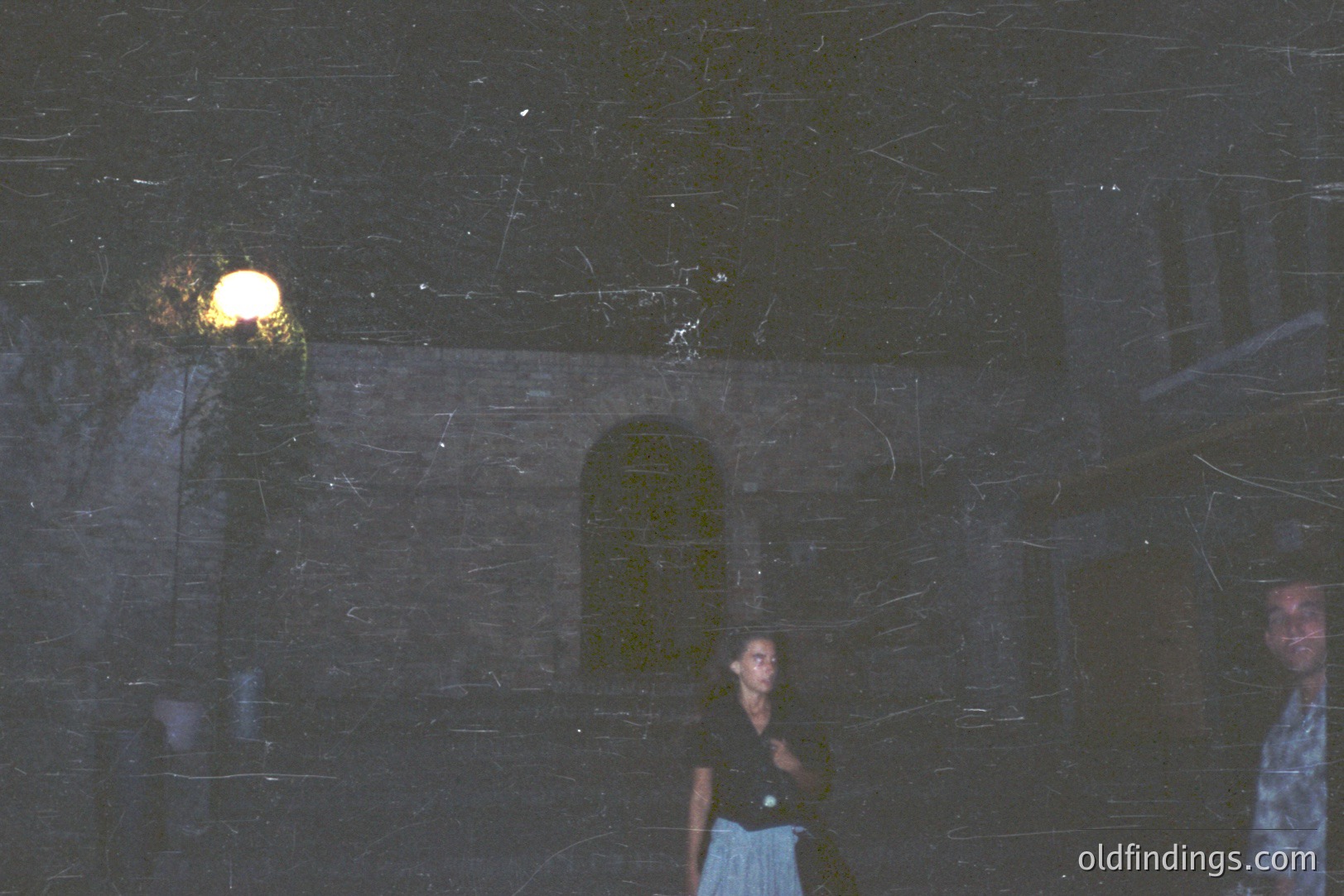 An exterior view featuring a woman and a man standing near a stone building with an arched doorway. The scene is dimly lit by a nearby lamppost, suggesting twilight or evening. The architecture evokes a Mediterranean or European style. Appears to be a candid snapshot, possibly from the 1970s. Potential stock photography value.