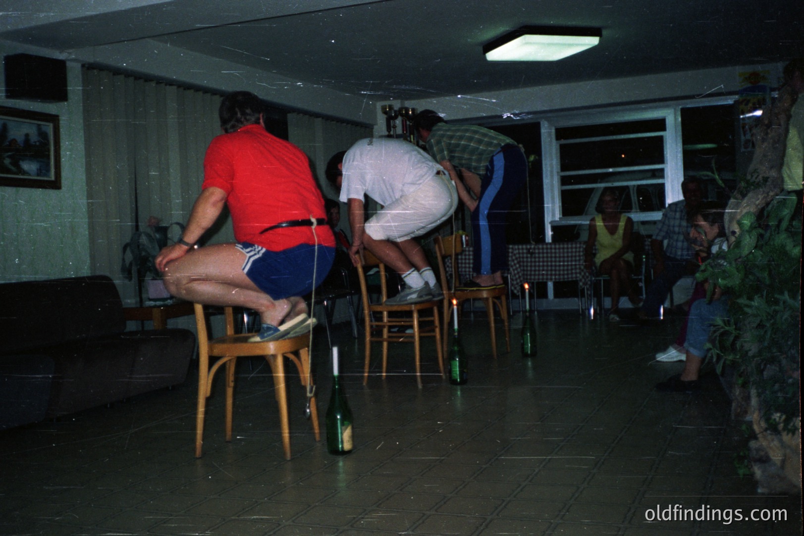 A group of people play "chair and bottle" in a dimly lit, modernist home. Three men, dressed in 1970s-era attire, balance on chairs around strategically placed bottles. Guests observe from a nearby counter. Window views hint at a seaside location. A striking snapshot of leisure and socializing.