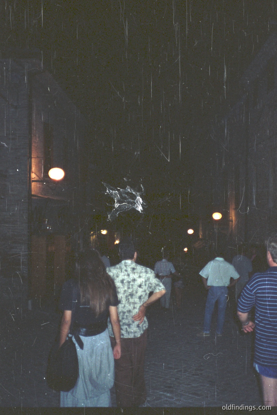 A group of people observes a suspended, ornate chandelier within a dark, arching passageway. The building features stone walls and lantern-like lights. Appears to be an interior courtyard or gallery. Likely 1980s era, potentially European. Valuable for design and architectural reference.
