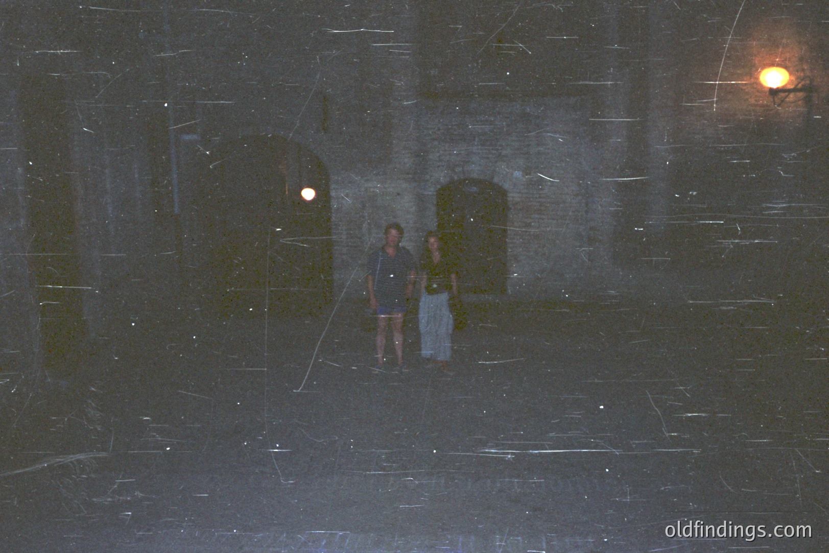 A couple walks along a cobblestone pathway within an ancient stone structure. Visible architecture includes arched doorways & a lit wall sconce. Likely 1970s/80s based on film grain & clothing styles. Unclear location, possibly Mediterranean. Shows aged aesthetic.