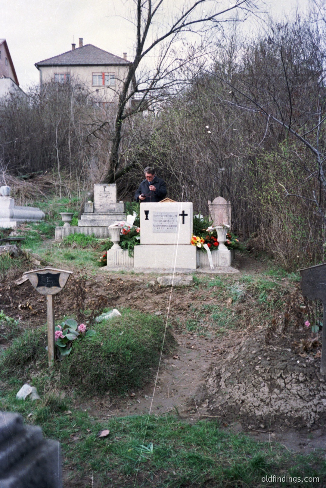 An elderly man stands before a memorial in a hillside cemetery, possibly Eastern Europe. Stone monuments and graves are visible amongst bare winter trees. The memorial features a cross and wreaths. A building is seen in the background. Likely 1980s-1990s.