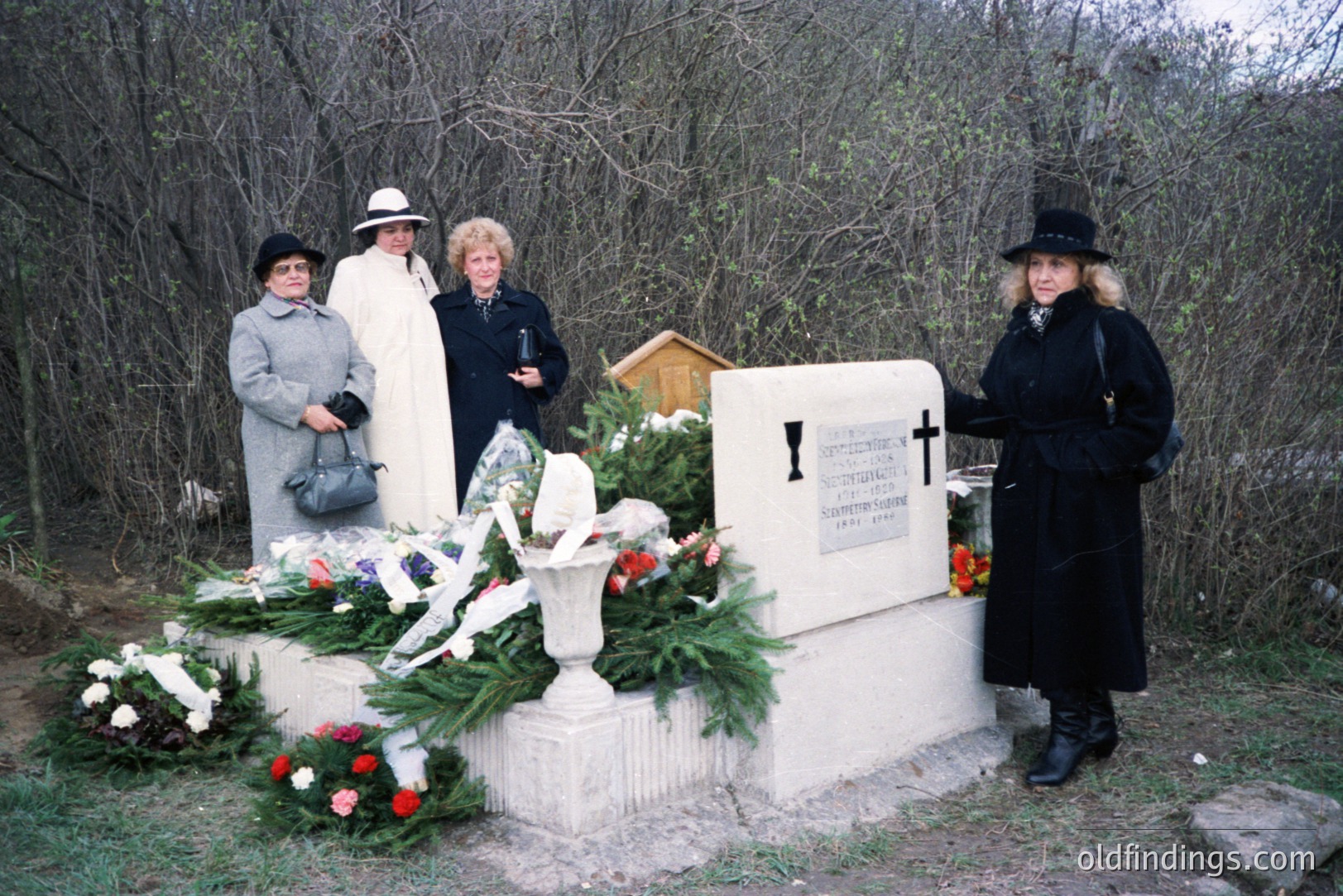 Four women in formal dress stand before a marble monument adorned with floral arrangements, possibly at a memorial or gravesite. Pine boughs and red roses add color. The setting appears to be a wooded area with a slight incline. Likely 1970s or 80s.