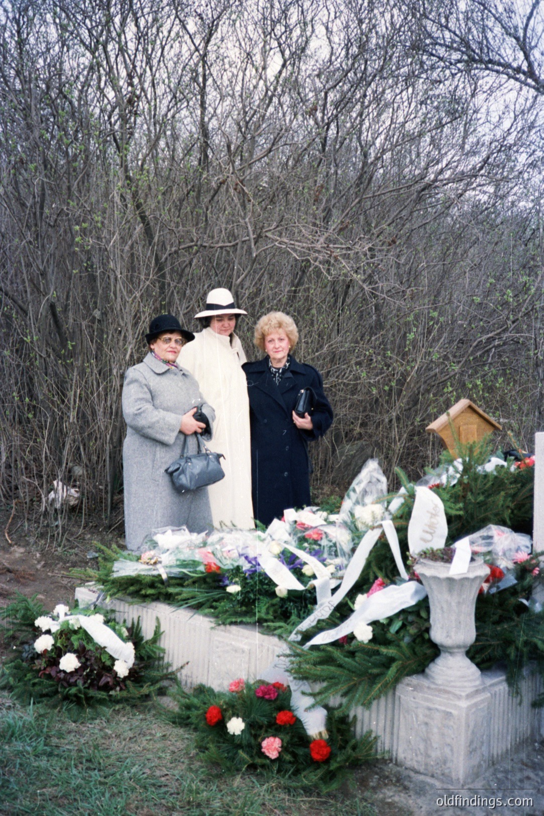 Three women stand before a stone monument heavily adorned with floral tributes. The women wear 1970s-era clothing and hats. A stone urn overflowed with flowers is central to the composition. Likely a memorial site or gravesite within a wooded, undeveloped area.