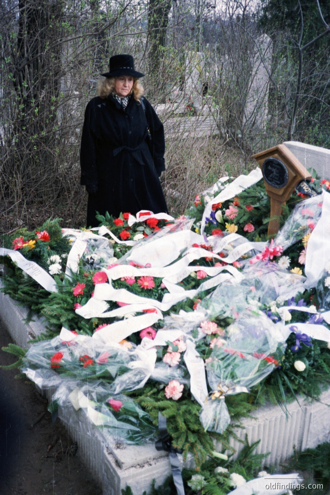 A woman in a dark coat and hat stands near a memorial, heavily adorned with floral tributes including roses, carnations, and ribbons. A plaque is visible on the memorial. Likely a gravesite, possibly commemorating an event or person of significance. 1970s-80s aesthetic.