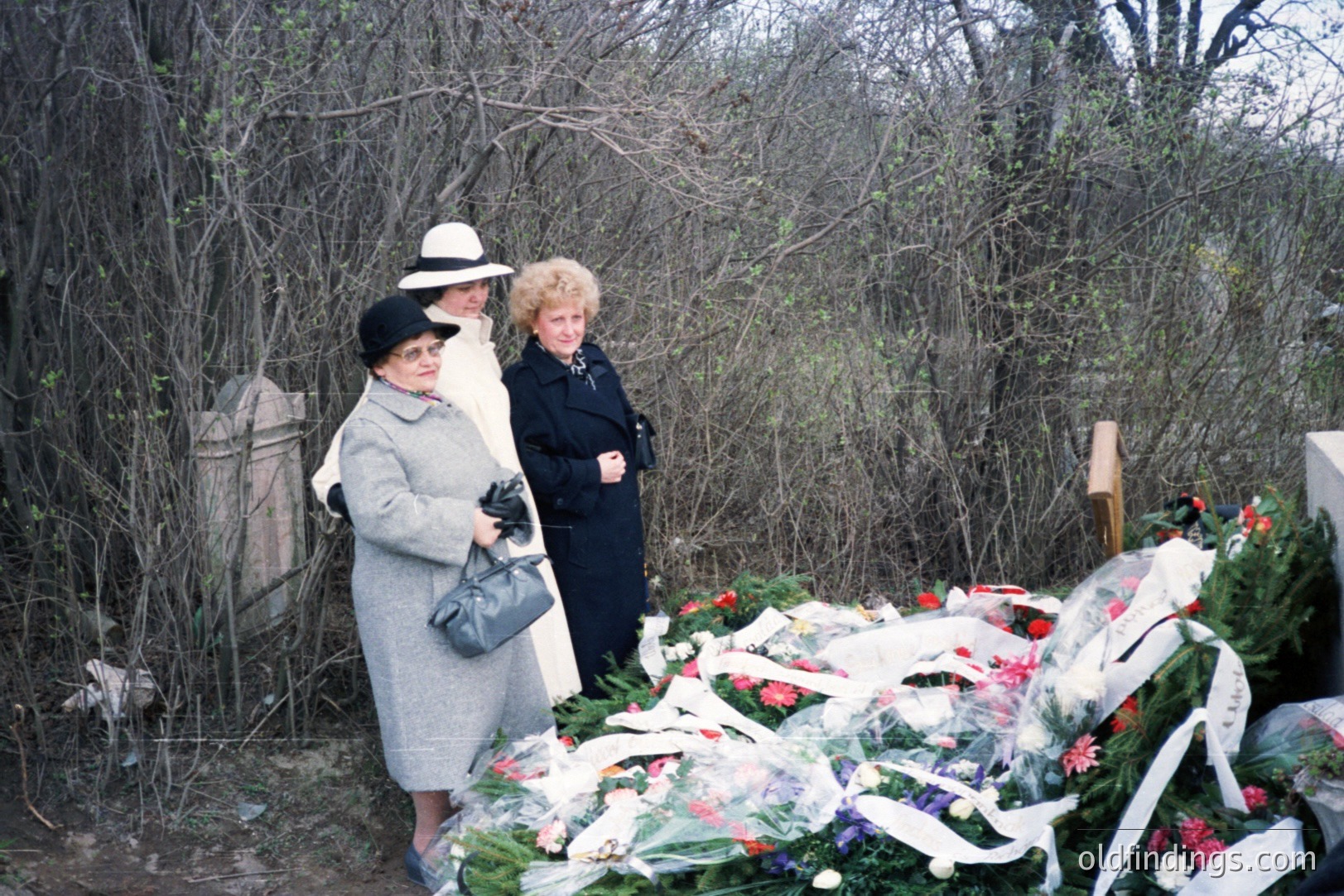 Three women in vintage attire stand beside a large arrangement of fresh flowers, likely at a gravesite or memorial. The women are dressed in 1960s/70s style coats and hats. Surrounding foliage suggests a rural or wooded area. The photograph’s tone and color palette evoke a sense of quiet remembrance and loss.