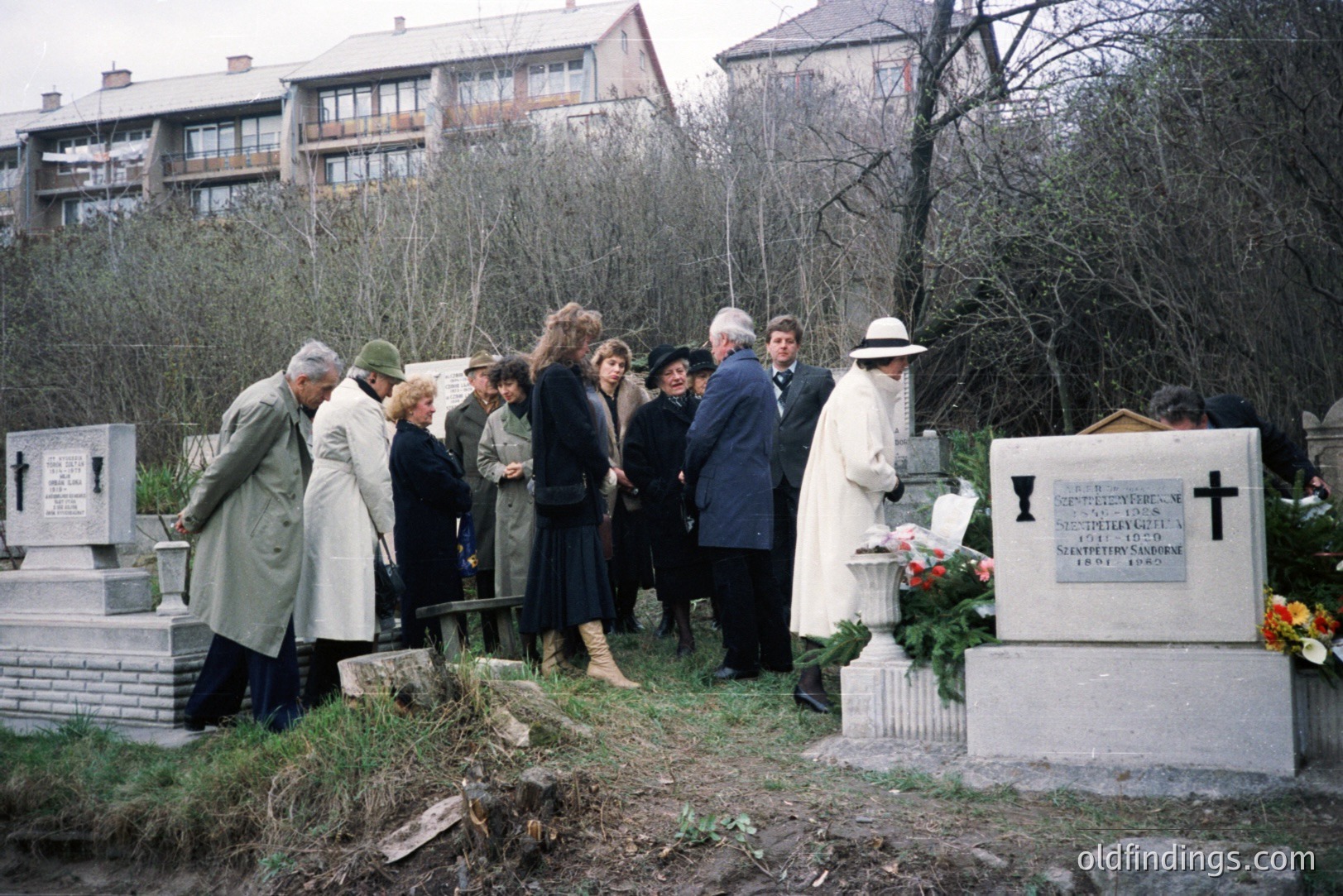Group gathers at a gravesite in a European cemetery. The scene features floral tributes, a stone cross, and a memorial plaque with Cyrillic inscription. Buildings are visible in the background. Likely 1970s or 80s, Eastern Europe. Reflective of post-war memorial culture.