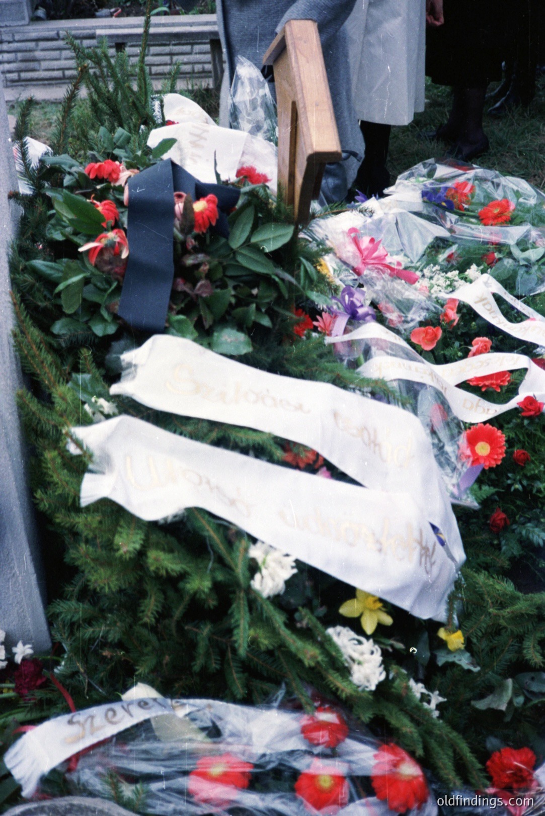 Elaborate floral tributes – reds, yellows, whites – sit atop a grave surrounded by pine branches. Handwritten messages on white ribbons adorn the arrangement. Likely memorial service; partial view of a person in dark clothing in the background. Appears to be a candid photo.