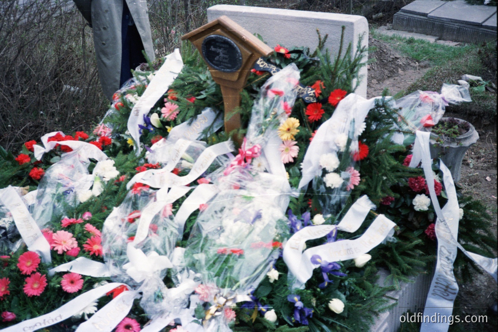 Elaborate floral arrangement with chrysanthemums, lilies, and ribbons adorns a gravestone. A small, wooden plaque is prominently displayed within the arrangement. The photo appears to be a snapshot, likely from the 1980s or 1990s, judging by the film quality and color palette.