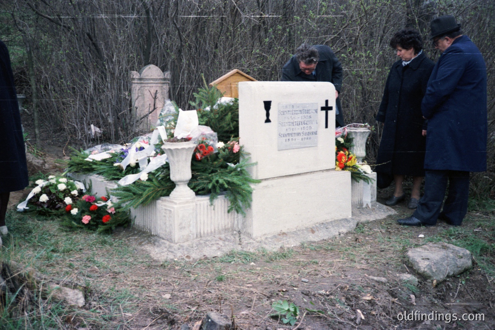 A solemn scene at a gravesite, featuring a large, ornate stone memorial adorned with fresh flower arrangements and a small, classical urn. Three figures, two men and a woman, are respectfully observing. The setting appears to be a rural cemetery. Likely Eastern Europe, late 20th century.