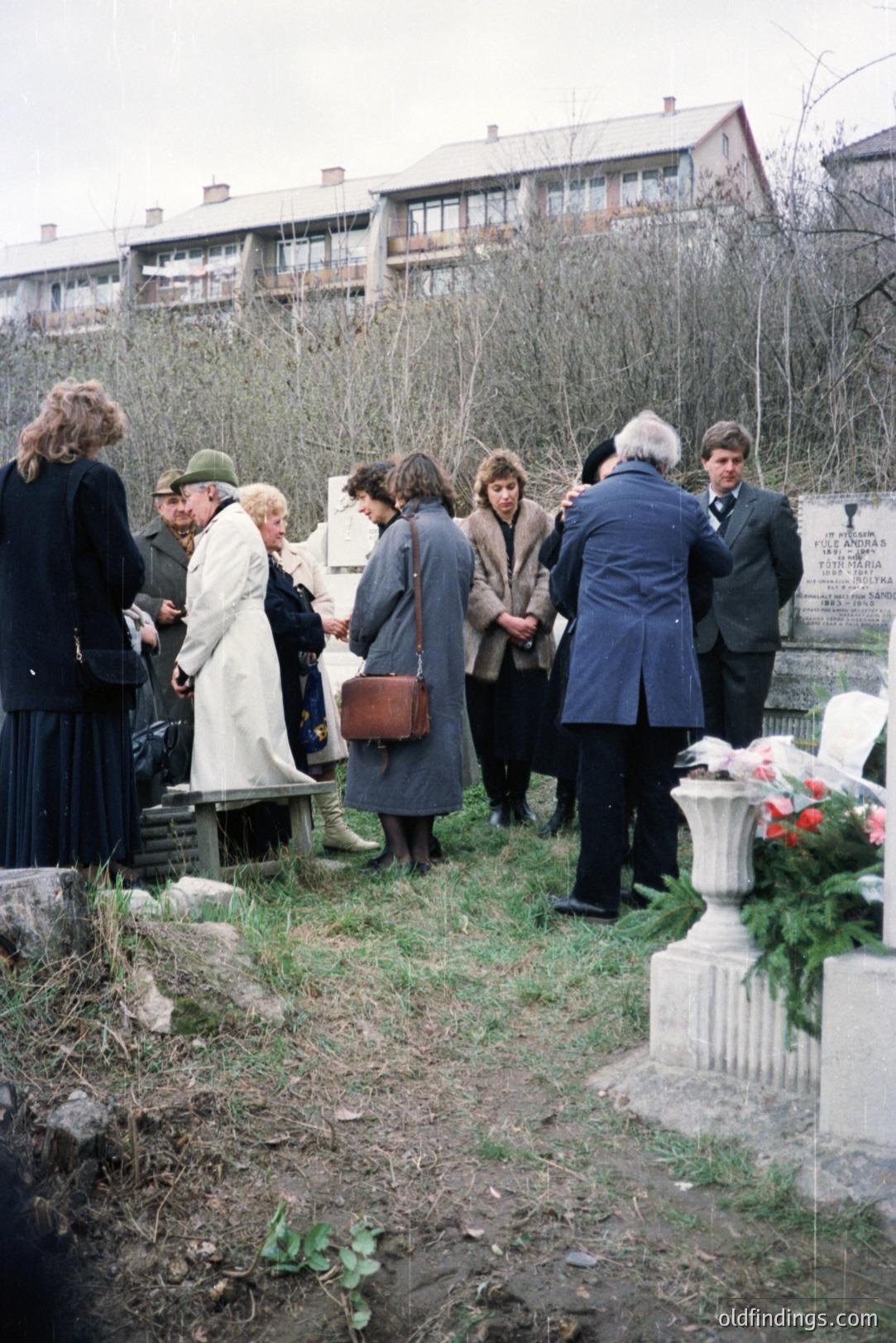 Group gathers around a memorial in a partially bare, muddy, landscaped area, framed by a brutalist apartment block. Figures dressed in 1970s-80s outerwear, including coats, hats, and a military-style jacket. Floral tributes adorn a stone urn. A somber, reflective atmosphere.