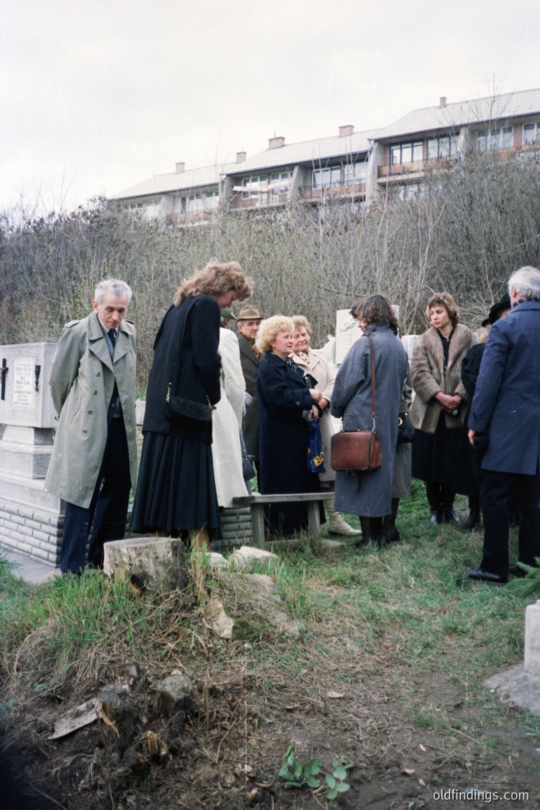 Group of people appears to be participating in a formal gathering, possibly a tour or memorial, in front of a brutalist-style apartment complex. Note the 1970s fashion and architecture. Likely Eastern Europe, perhaps Bulgaria. Location obscured by foliage. Limited natural light.