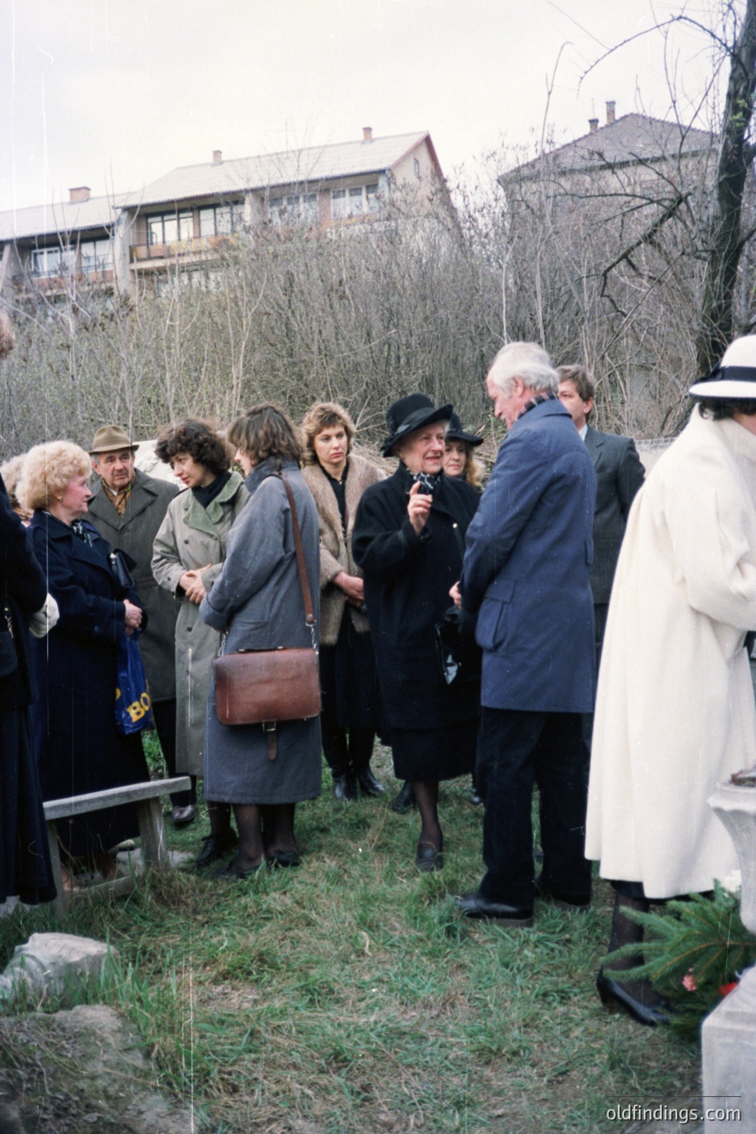 Group gathers for a somber outdoor memorial or ceremony. People are formally dressed in coats, hats, and scarves; likely a remembrance event. Background shows a modest, multi-story apartment building. Appears to be a public space, possibly near a gravesite. Estimated 1970s-1980s.