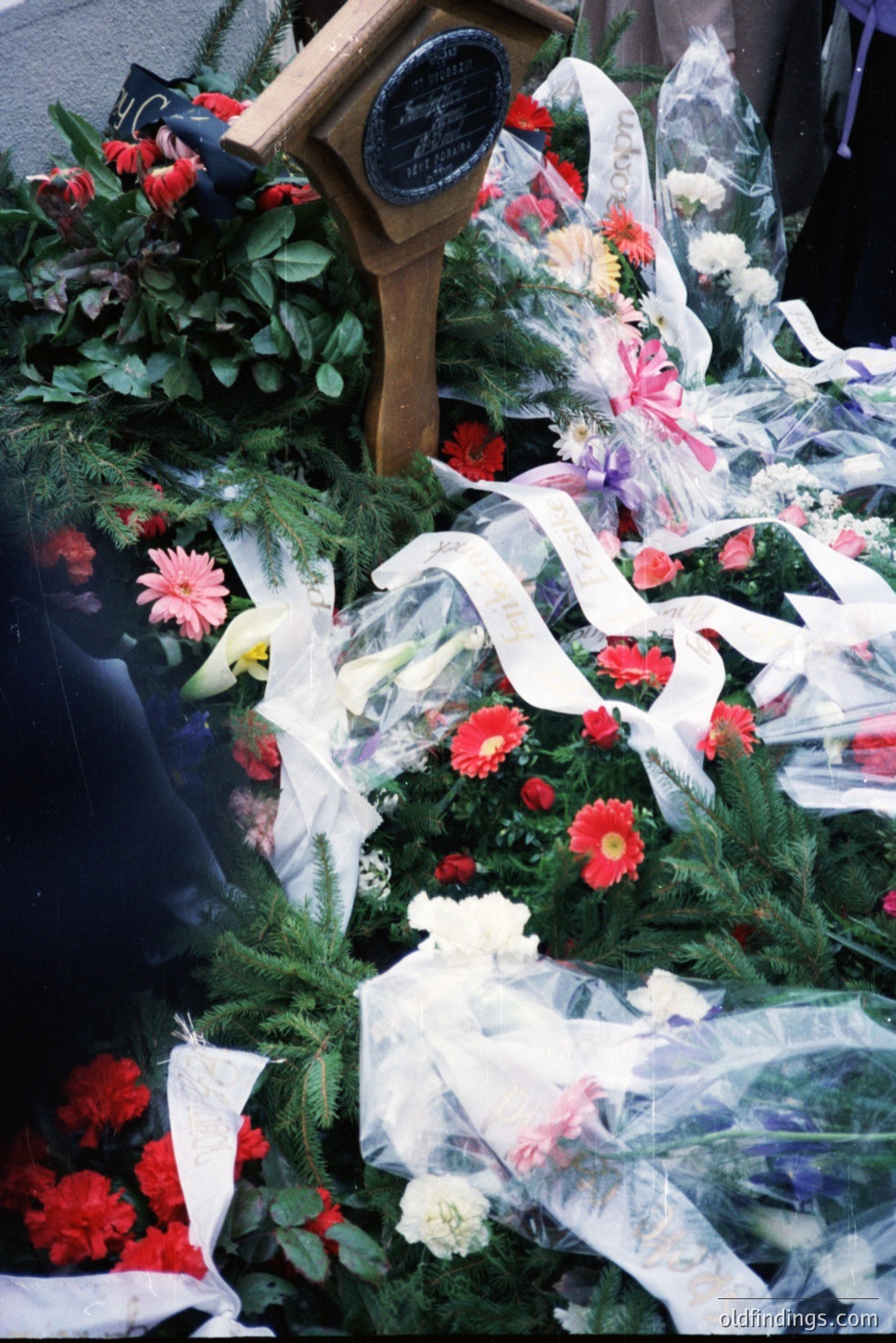 A memorial display features vibrant red Gerbera daisies, white roses, and ferns, accented by flowing white ribbons. A dark plaque is partially visible. Likely a funeral or memorial service scene, circa 1980s/90s judging by the floral arrangement style.