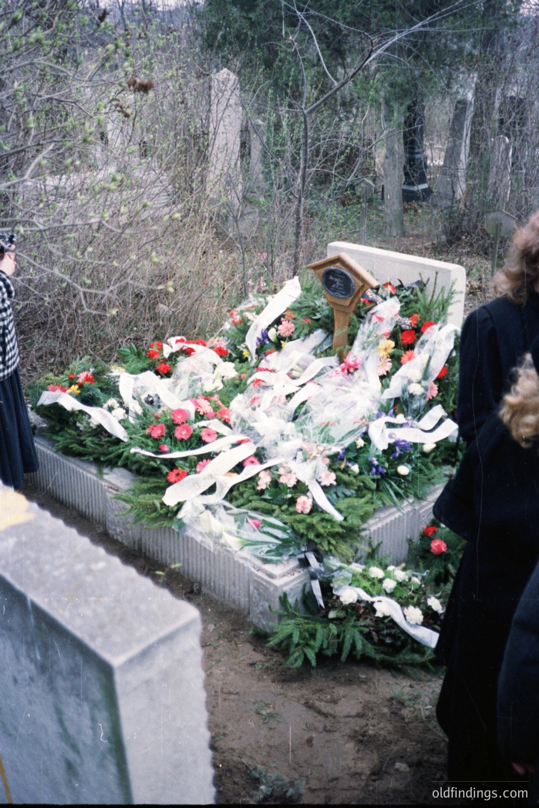 Overgrown concrete memorial platform, heavily adorned with floral tributes and ribbons. The platform appears to be within a wooded area, likely a cemetery or memorial site. Partial view of two figures in dark clothing are visible in foreground. Appears to be a late 20th-century scene, possibly 1980s.