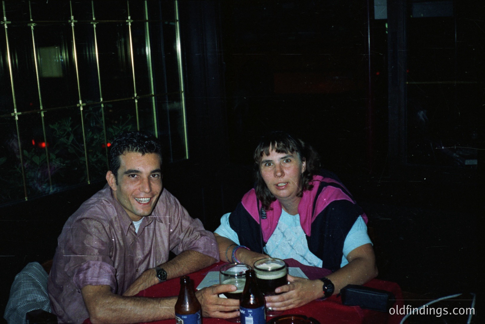 Two individuals sit at a table with drinks during nighttime; a beer and bottled beverages visible. The man wears a patterned button-down shirt. The woman’s attire includes a white sweater with pink and purple accents. Likely a candid snapshot from the 1980s or 1990s.