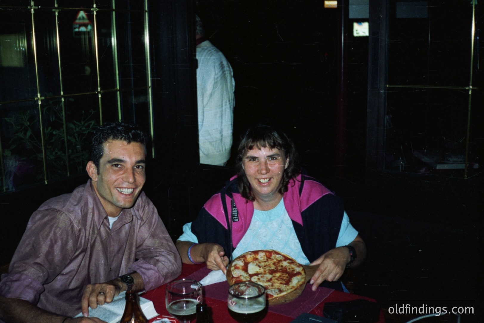Two people seated at a table with a pizza, likely outdoors at night. Man in a short-sleeve shirt smiles directly at the camera. Woman wears a color-block tracksuit, holding a fork. A glass of red liquid & bottle are visible.