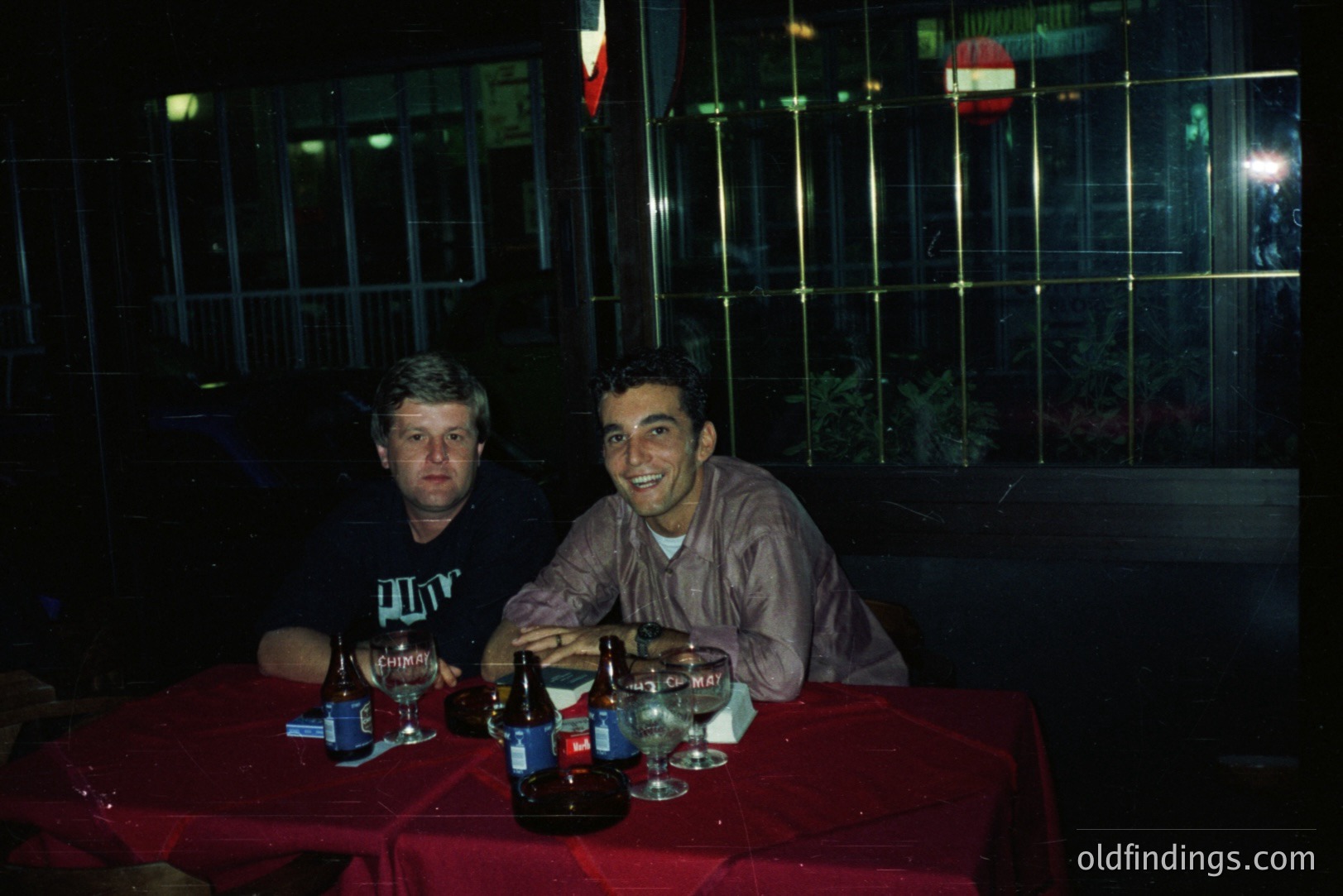 Two men seated at a table covered with a vibrant red tablecloth, appear to be enjoying drinks, likely beer. The setting is outdoors, possibly a patio or balcony, with a metal railing visible in the background. Photo likely taken in the 1990s based on clothing and color palette. Casual, candid moment.