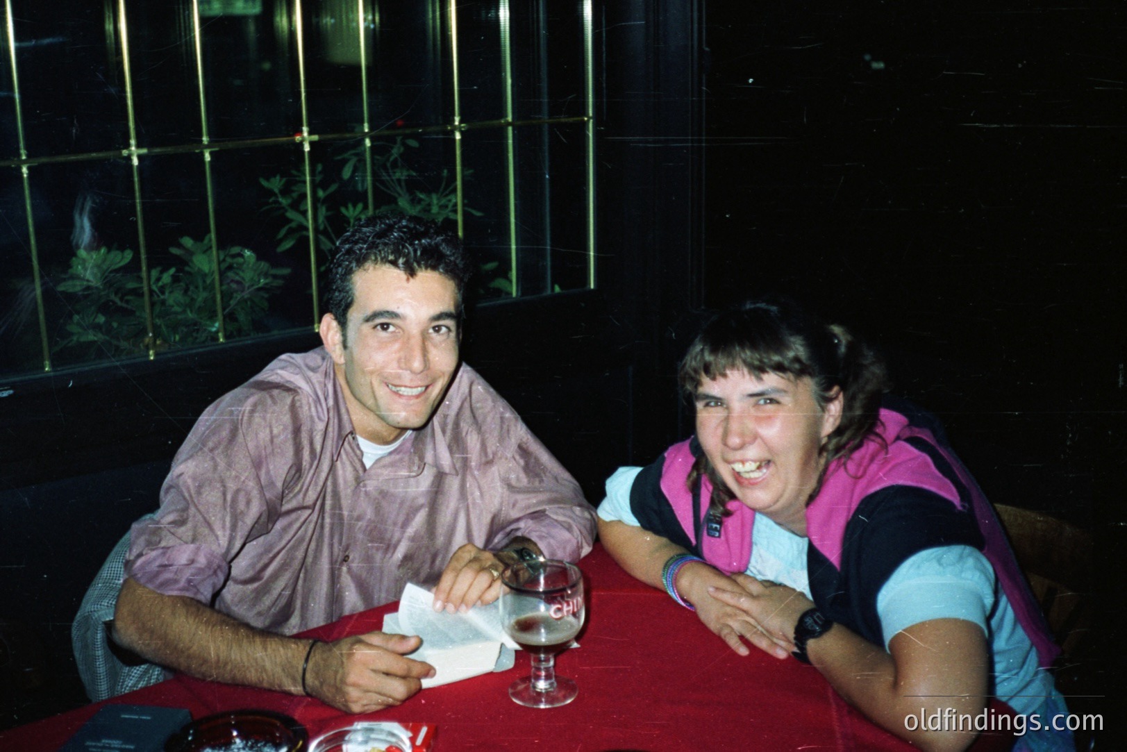 Two young adults seated at a table covered with a vibrant red tablecloth smile for the camera. A glass of beer sits on the table between them. The image appears to be a snapshot, likely taken in the 1990s, given the clothing and photographic style. A dark background with foliage suggests an outdoor or semi-outdoor setting.