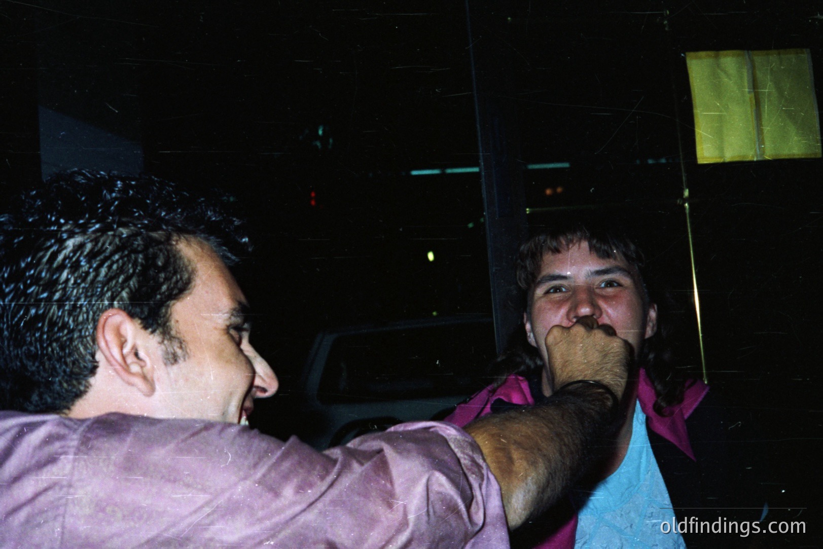 Two men share a playful moment in a dimly lit, possibly outdoor setting. One man, in a purple shirt, smiles while appearing to gently restrain the other, who wears a pink shirt and has his face pressed against the first man’s arm. Appears to be a casual snapshot.