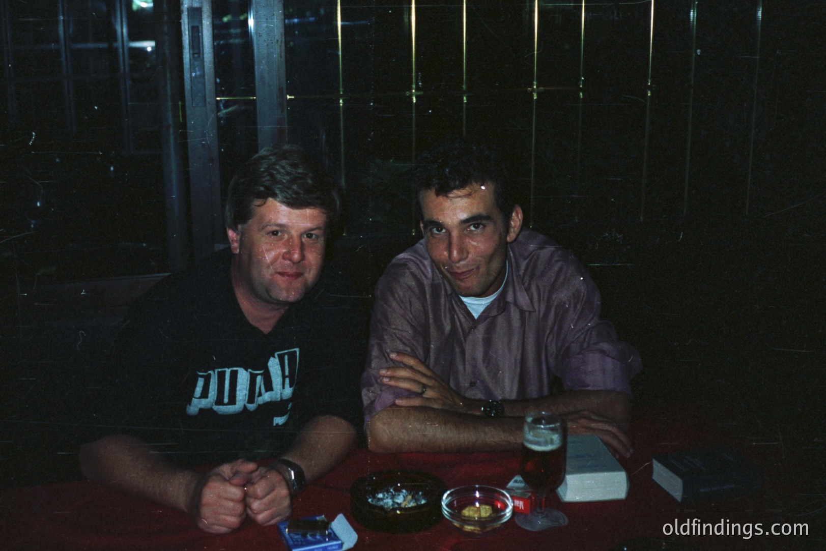 Two men sit at a table in a dimly lit, likely bar or pub setting. One wears a black Puma graphic tee, the other a purple shirt with his arms crossed. Visible are glasses, a beer, and an ashtray with cigarettes. Likely late 1990s/early 2000s casual scene.