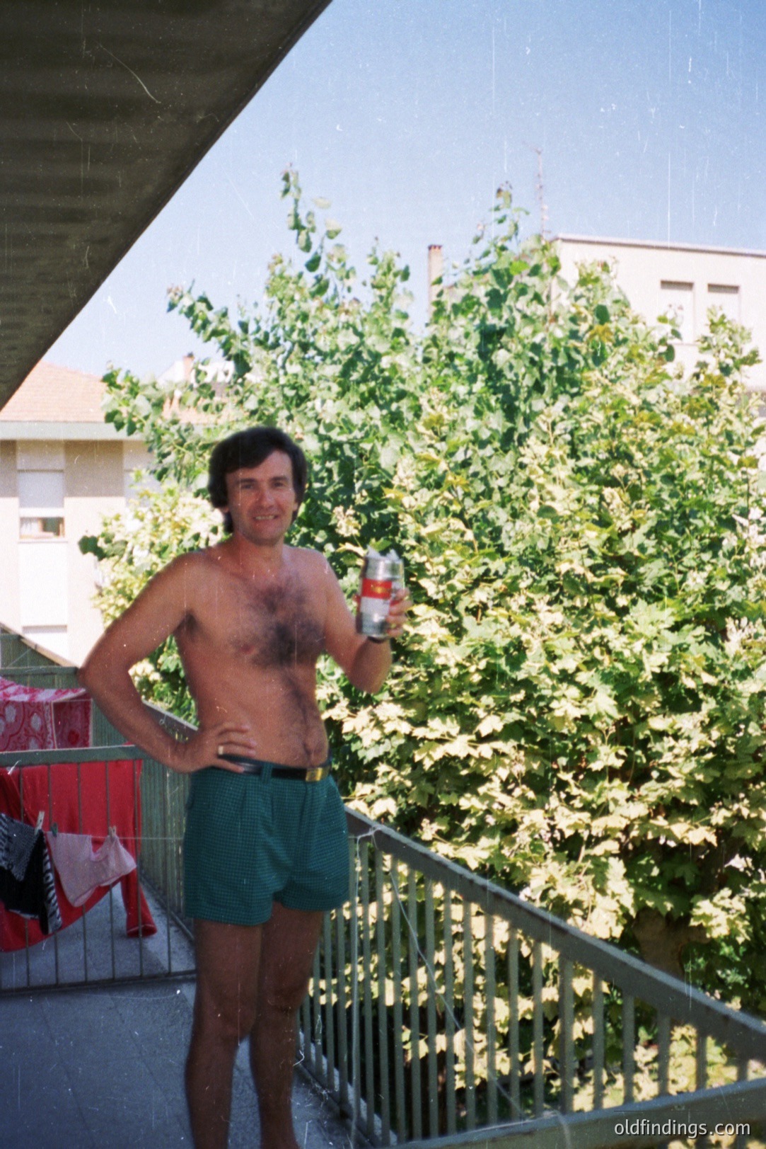 Man stands on a balcony, holding a beverage, outdoors. Green patterned shorts, visible chest hair. Balcony features metal railing & red chairs. Likely a vacation or leisure scene. Appears to be 1970s or 1980s film photography. Could be valuable for stock or design.