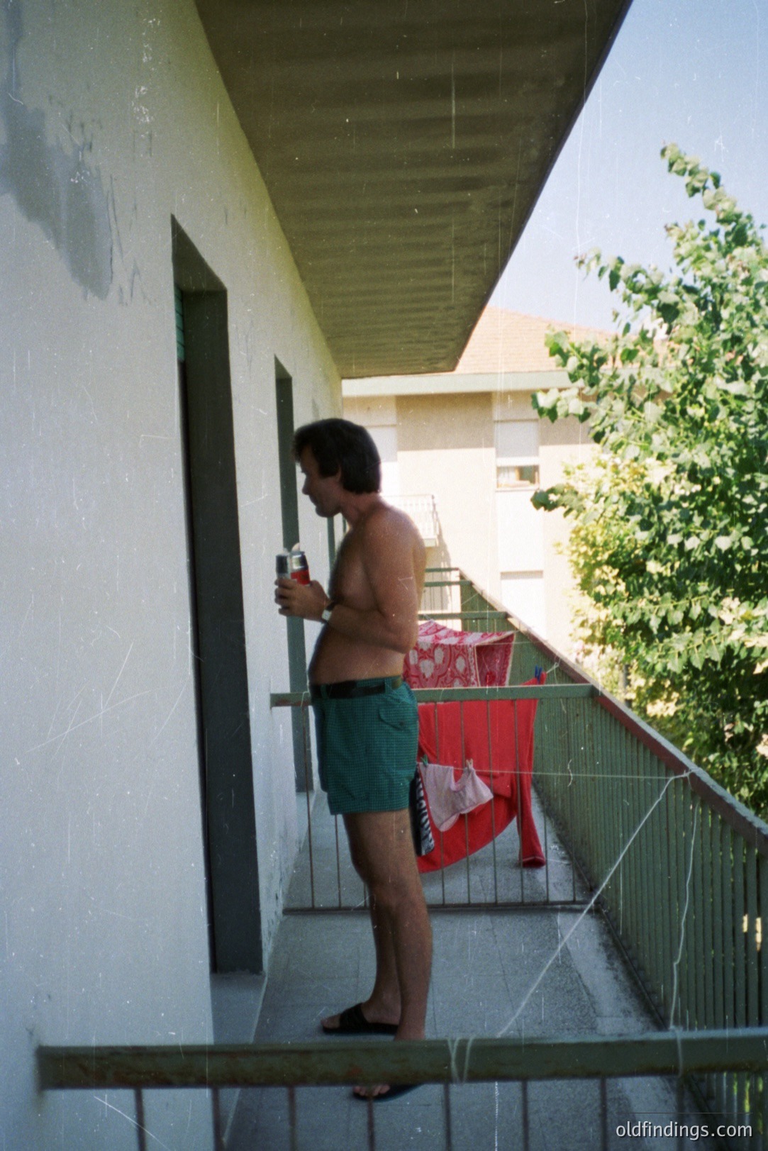 A man stands on a balcony, holding a can, appearing relaxed in patterned swim trunks and flip-flops. Laundry hangs to dry on a rack. Architectural style suggests mid-century Eastern European apartment complex. Likely 1970s. Scene evokes leisure and vacation time.