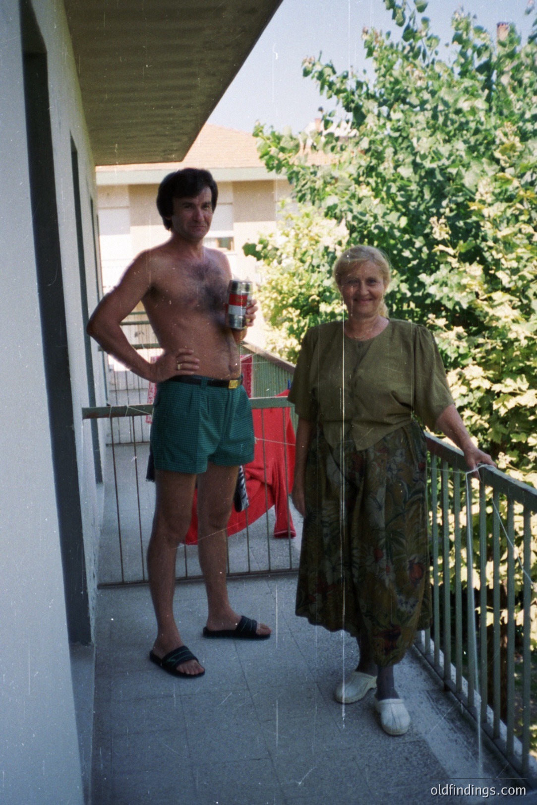 A man and older woman stand on a balcony; the man is shirtless, wearing swim trunks & sandals, holding a drink. The woman wears a floral-print dress and slippers. Likely a 1970s holiday snapshot, showing casual vacation style. Balcony features metal railing and exterior view.