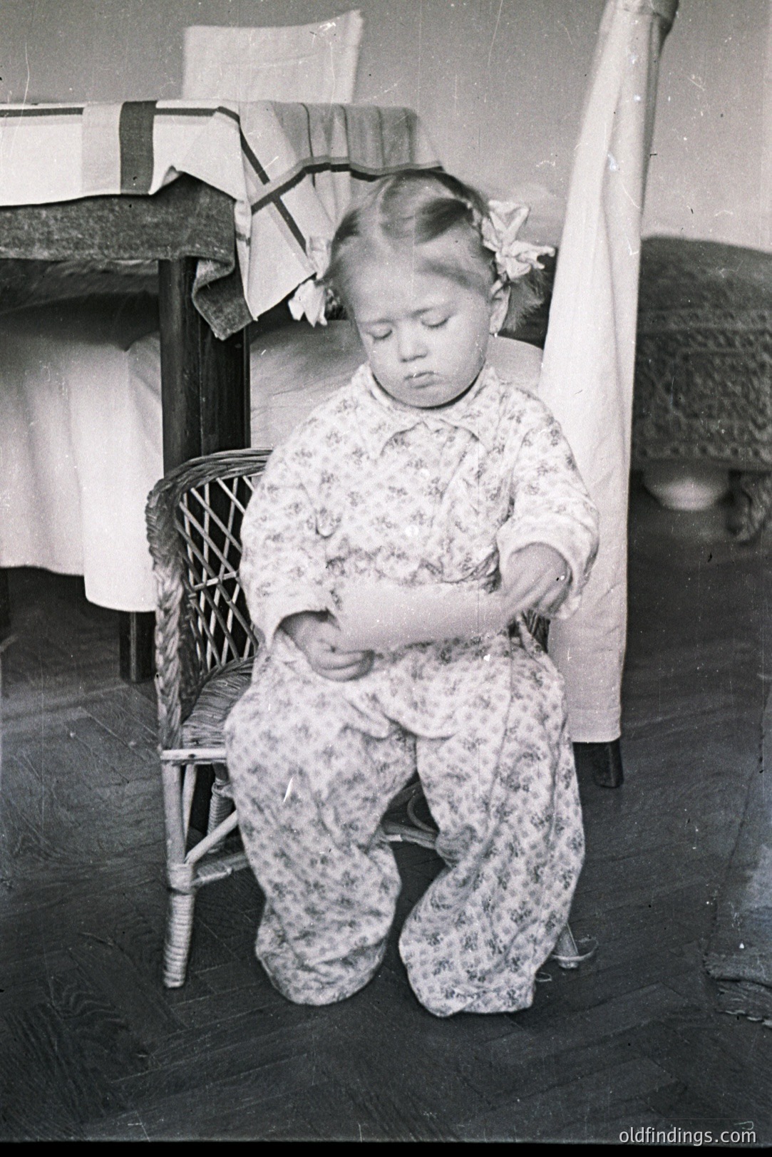 A young girl, likely toddler age, kneels on a wood floor before a rattan chair. She wears a patterned floral dress and a flower in her hair. The setting suggests an interior, possibly a bedroom with draped curtains visible. Circa 1910-1930, likely a family snapshot.