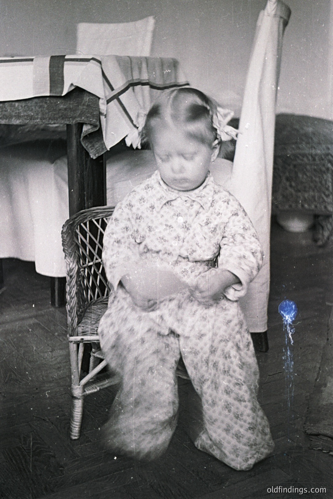 A young child seated on a wicker chair, posed formally, wears a floral print dress and ribbon in their hair. The backdrop suggests a domestic interior, potentially a bedroom with a bed and curtains. Likely a studio or candid family portrait from the 1930s-1950s. Delicate and evocative.