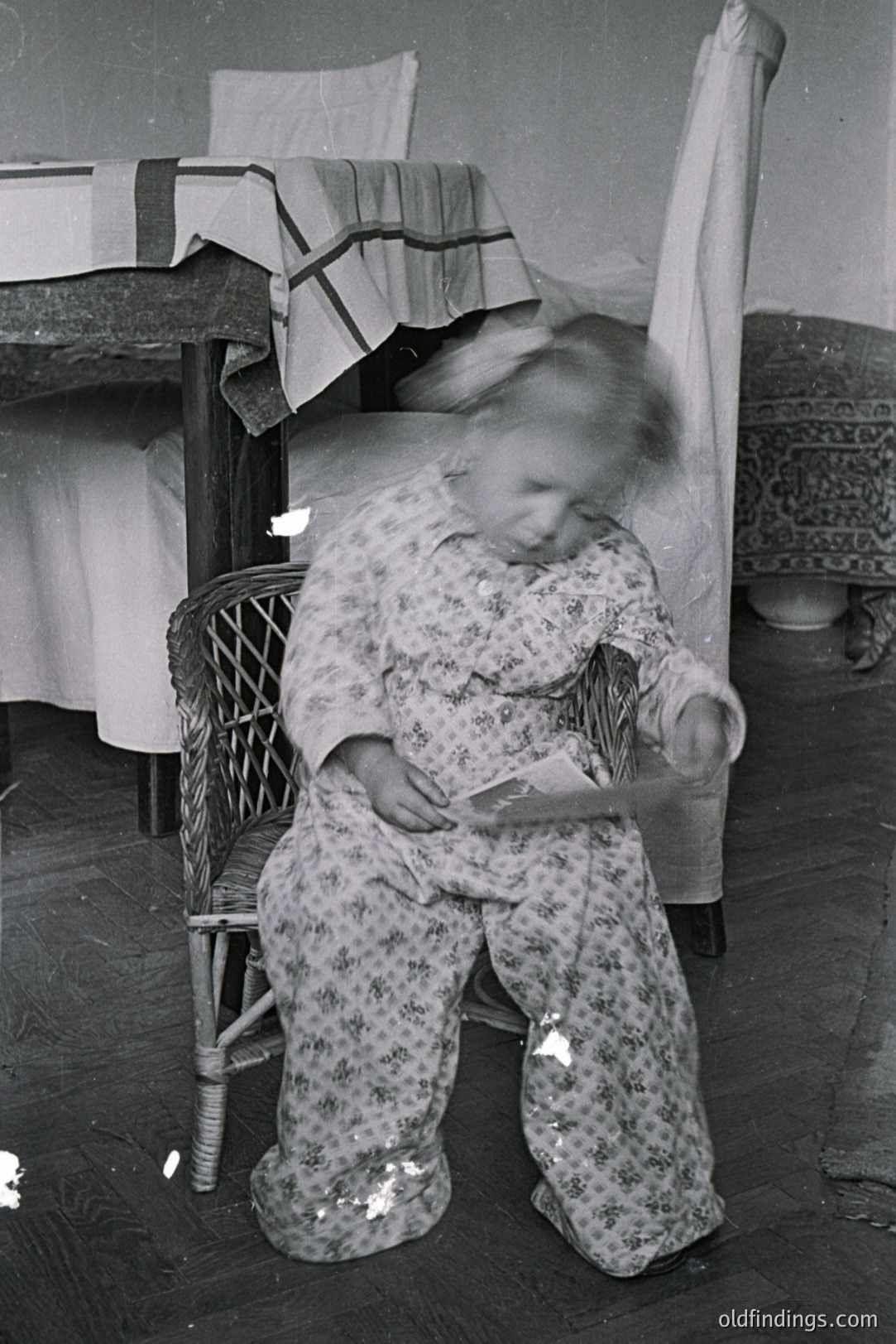 A young child, blurred with motion, sits in a wicker chair holding a small paper or booklet. The scene features a patterned bed with a ladder and hardwood floors, suggesting a residential interior. Likely a candid moment, possibly captured in the mid-20th century. Documentation of childhood activities.