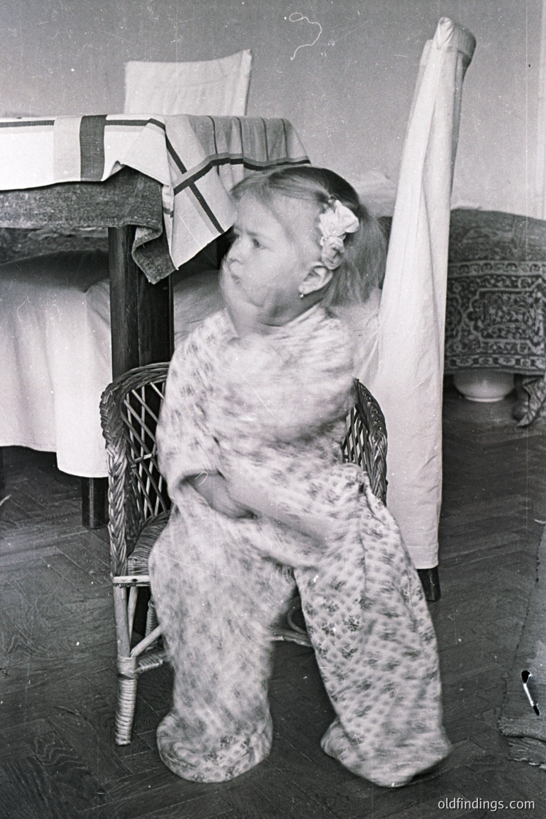 A young girl sits in a wicker chair, wearing a floral patterned robe with a matching headband. The room features a bed with a patterned comforter and a patterned rug. Likely a domestic interior, c. 1940s-1960s. Potential stock reference for vintage childhood fashion.