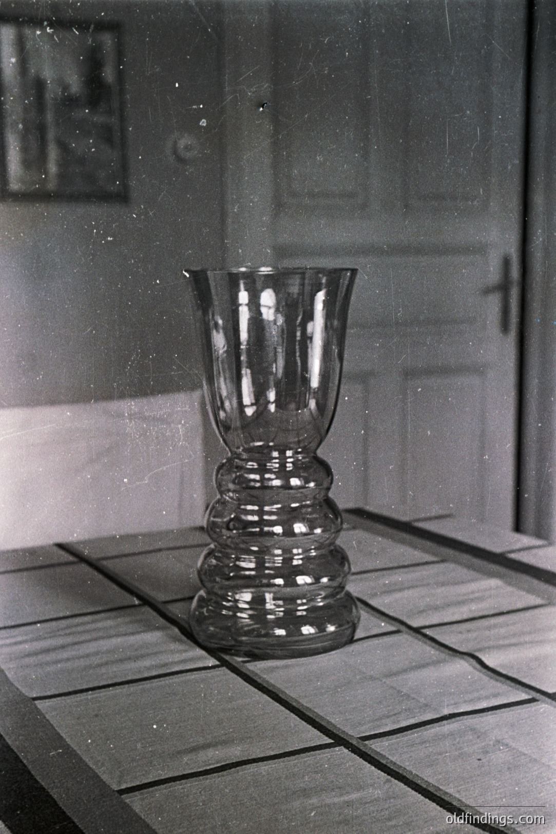 A tall, intricately-designed glass vase stands on a tiled surface. The vase features a flared top and a stepped, bulbous body with stacked rings. Interior door panels and a framed artwork are visible in the background. Likely a studio still life, potentially from the mid-20th century.